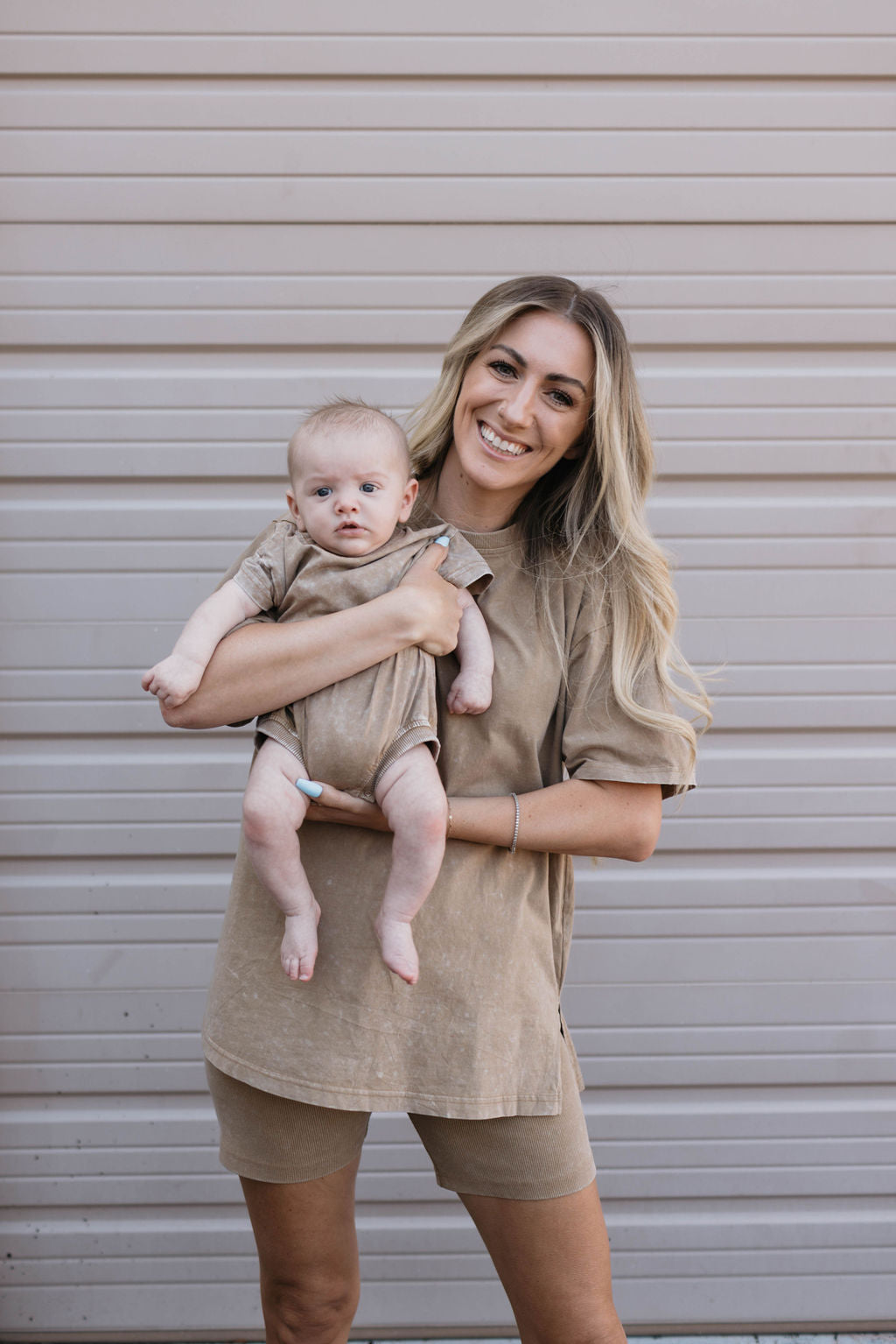 A smiling woman with long blonde hair is holding a baby wearing an adorable Forever French Baby onesie. Both are dressed in matching beige outfits and standing in front of a light grey garage door. The woman is wearing the Women's Short Set | Vintage Washed Espresso from the brand Forever French Baby.