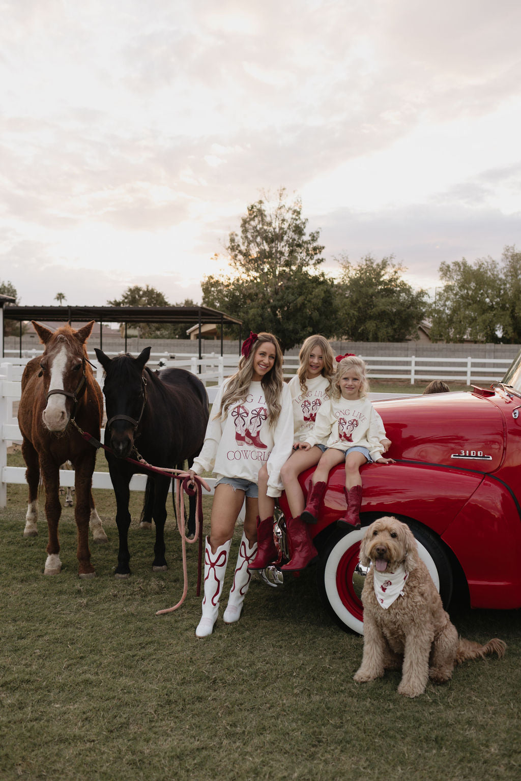 A woman and two young girls dressed in matching "Cowgirl Christmas" outfits stand beside a red vintage truck, while their horses and dog proudly display stylish Bamboo Bandanas from lolo webb. This picturesque ranch scene, with its white fence and surrounding trees, exudes the charm of a holiday postcard moment.