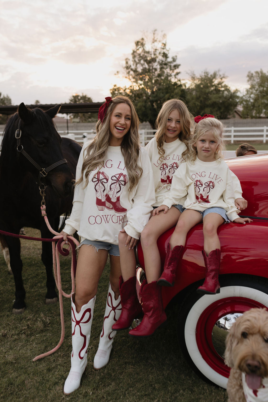 A woman and two children wearing cotton "Adult Crewneck | Long Live Cowgirls" shirts from lolo webb, along with denim shorts, pose beside a vintage red car and a black horse. Their outfits feature red and white colors, including cowboy boots, while a dog plays in the foreground. This charming scene takes place on a ranch.