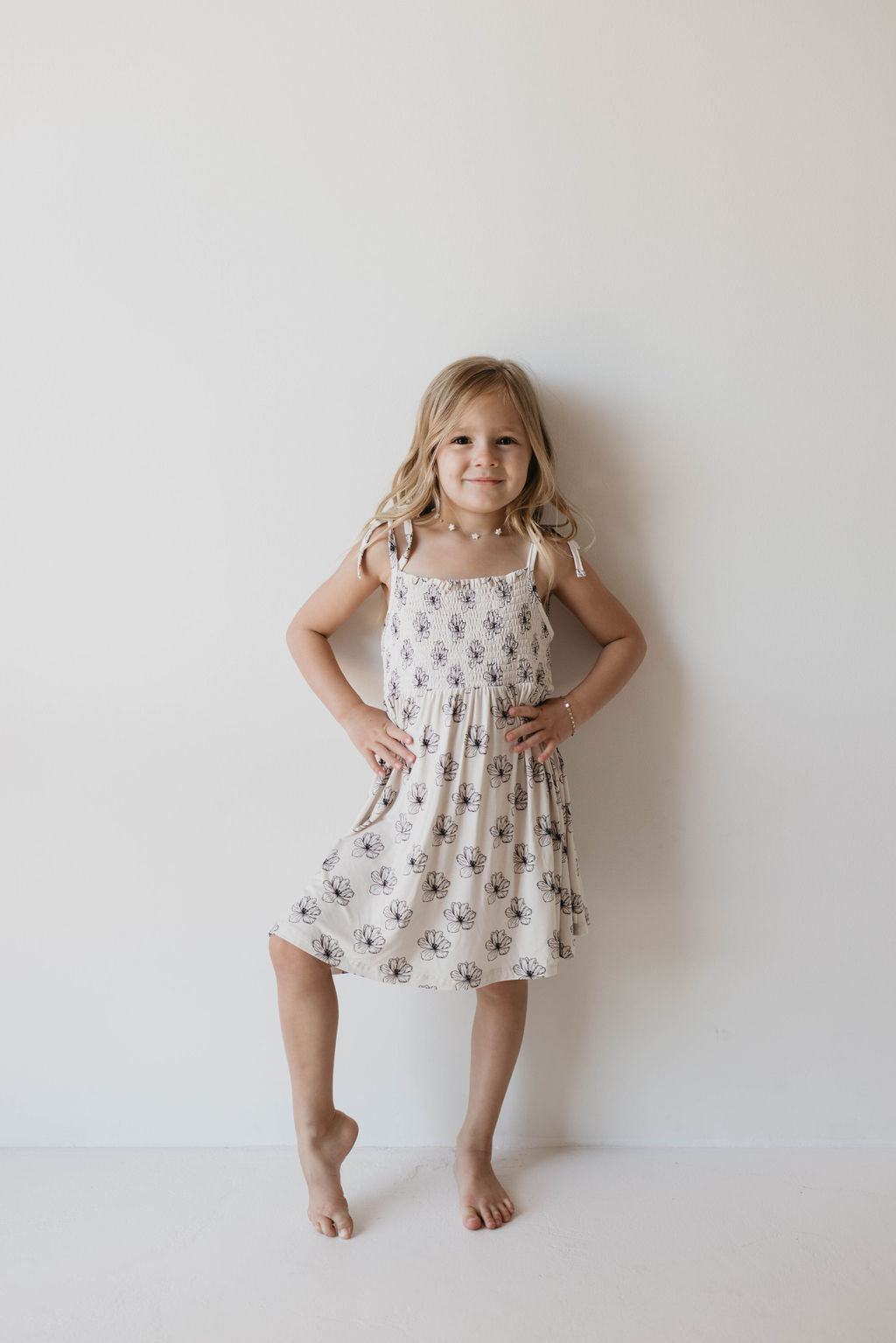 A young girl stands against a plain white wall, smiling with her hands on her hips. She is wearing the Bamboo Tie Top Dress in Desert Bloom by forever french baby and is barefoot. Her blonde hair is styled in loose waves, completing her effortlessly fashion-forward look.