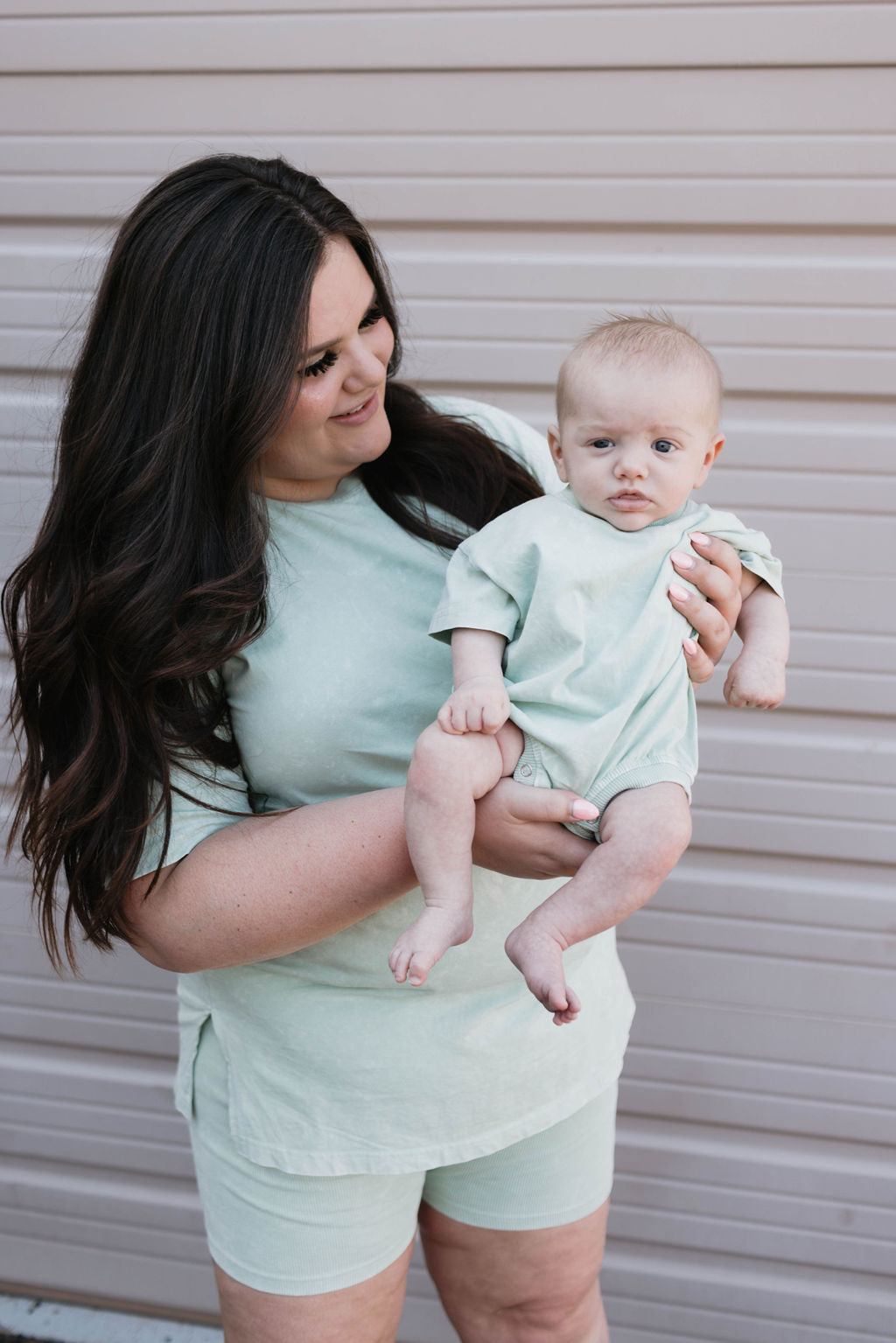 A woman with long, dark hair is holding a baby. Both dressed in the charming shades of the Women's Short Set in Vintage Washed Spearmint from forever french baby, they smile warmly at each other. They are standing in front of a beige garage door, creating a timeless moment captured Forever French Baby style.