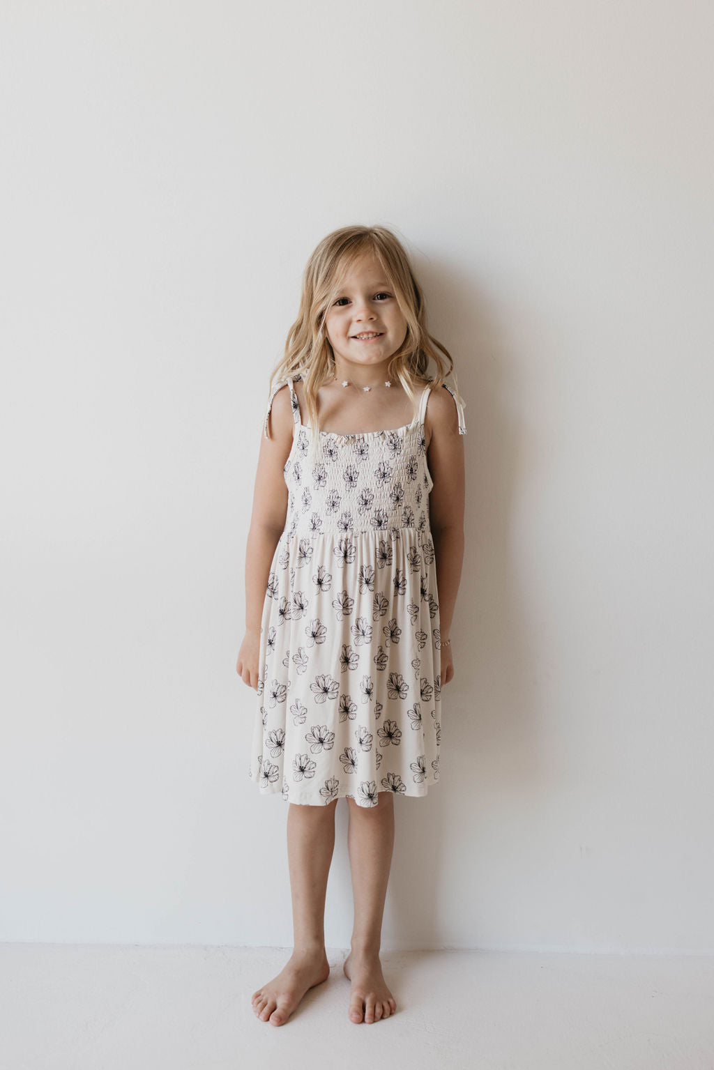 A young girl with long, blonde hair stands barefoot against a plain white wall, smiling. She is wearing the sleeveless Bamboo Tie Top Dress in Desert Bloom with floral designs from forever french baby.