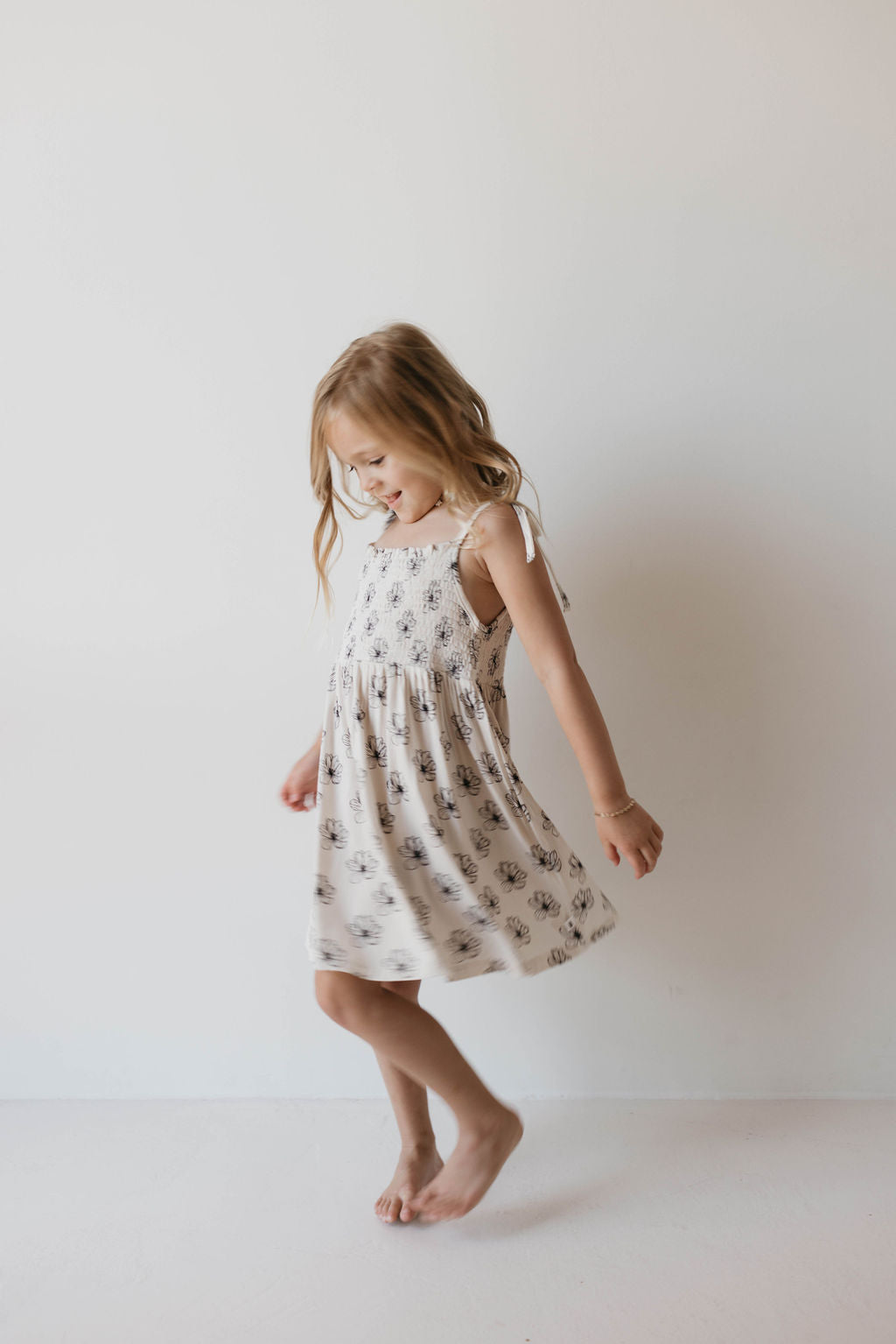 A young girl with long, wavy, blonde hair twirls joyfully in a light-colored, patterned sleeveless Bamboo Tie Top Dress from the forever french baby brand. She is barefoot and has a big smile on her face. The plain white background adds to the simplicity and focus on her joyful expression and fashion-forward movement in the Desert Bloom dress.