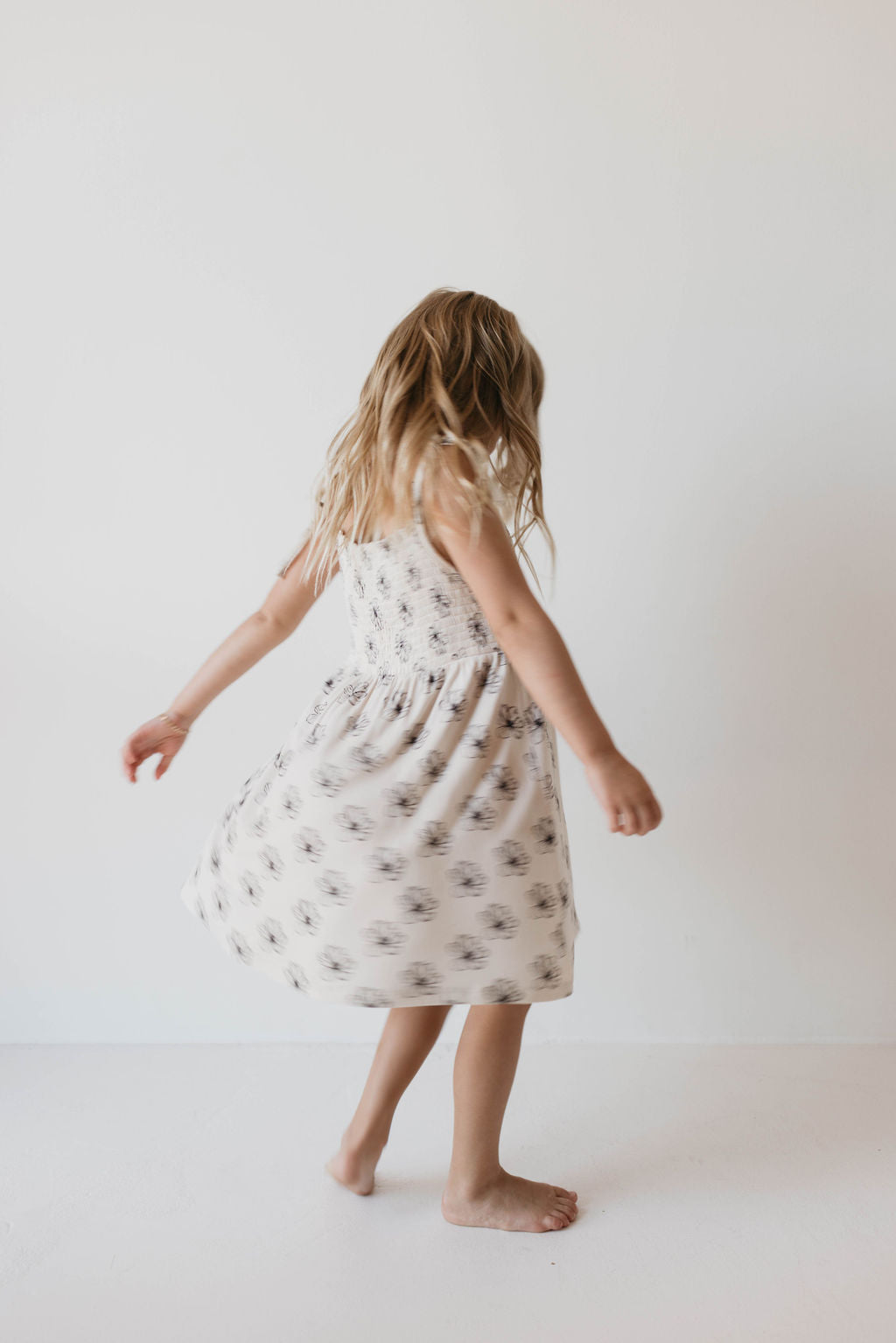 Against a plain white background, a young, fashion-forward child with long blonde hair is twirling barefoot in the "Bamboo Tie Top Dress | Desert Bloom" by forever french baby, featuring a white fabric patterned with small black flowers.