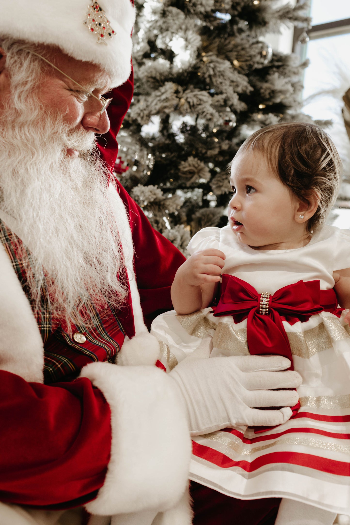 A person dressed as Santa Claus holds a young child in their lap. The child, adorned in a stunning white dress with red and gold accents from Forever French Baby, looks intently at Santa. They are near a beautifully decorated Christmas tree, with lights twinkling in the background; the perfect scene captured by Amanda Riley Photos for their "Santa Photos | Forever French Baby x Amanda Riley Photos" collection.