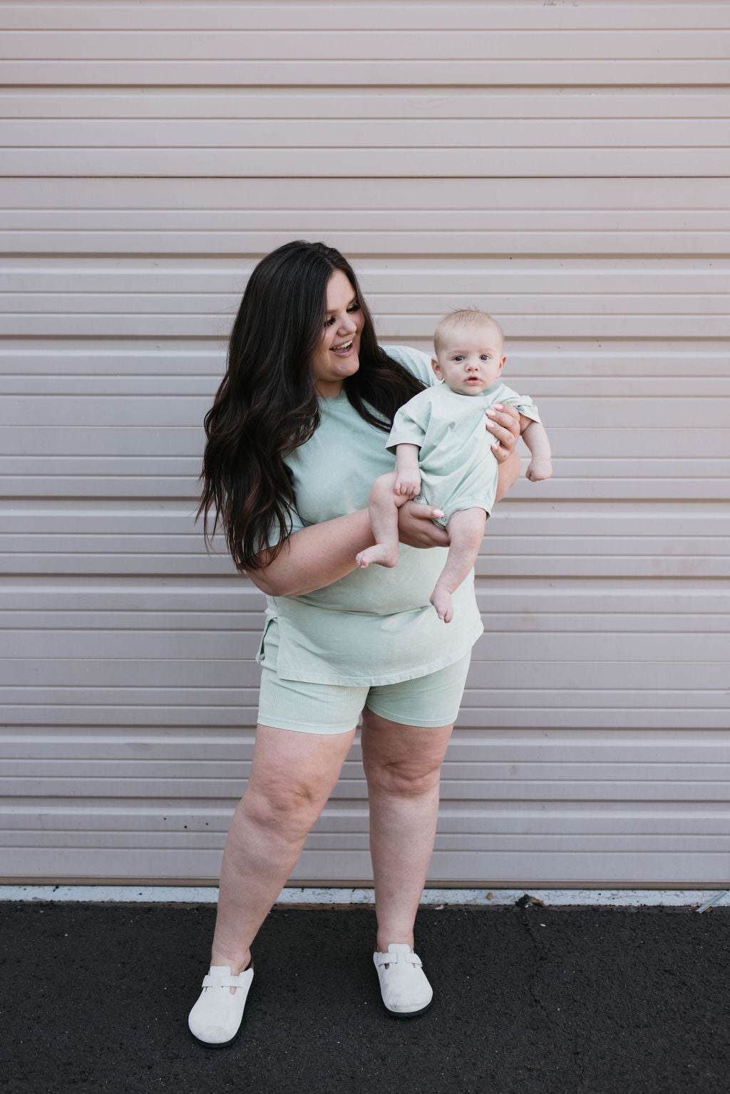 A woman with long dark hair, dressed in the Women's Short Set in Vintage Washed Spearmint, holds a baby who is wearing a matching Forever French Baby outfit. They stand in front of a corrugated metal backdrop. The woman smiles as she gazes at the baby.