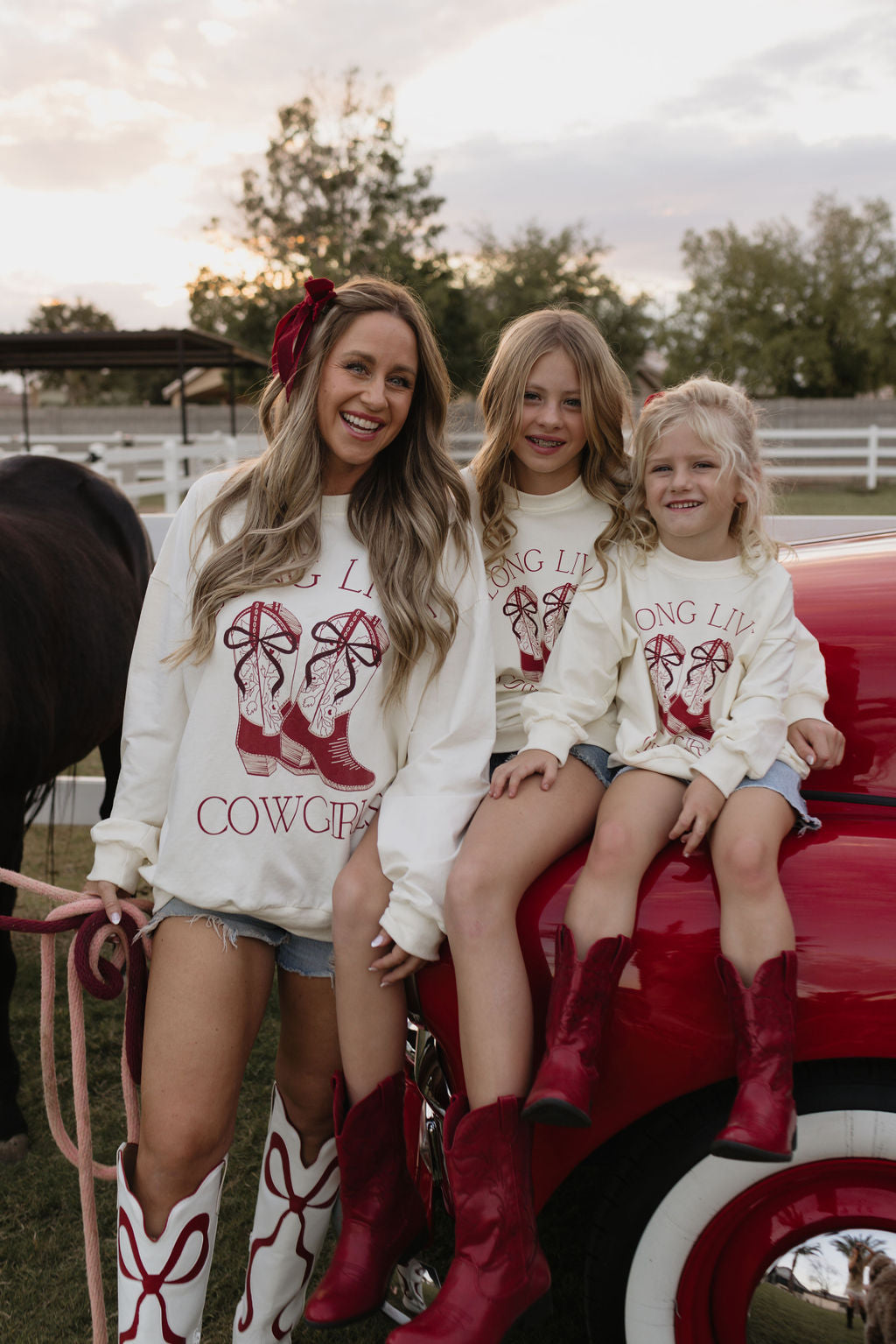 Three smiling people are wearing lolo webb's "Adult Crewneck | Long Live Cowgirls" cotton shirts and red boots. Two kids, in matching children's versions, sit on a red truck, while the adult stands nearby holding a rope. A horse and a fence provide the charming backdrop for this scene.