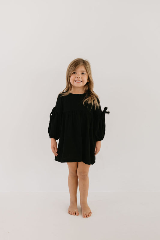 A young girl with long hair stands barefoot on a white background, wearing the stylish "Aria" dress from forever french baby, featuring long sleeves and bows. She smiles while facing forward, radiating elegance.