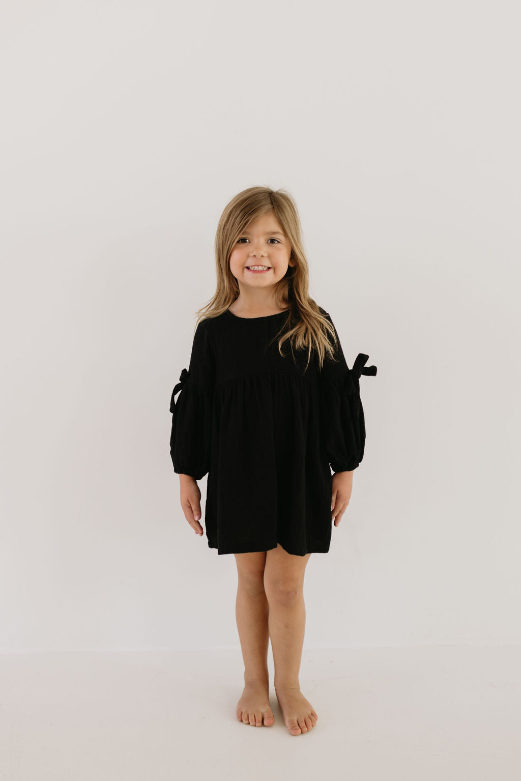 A young girl with long hair is smiling, standing barefoot against a neutral background. She wears the Child Dress | Aria by forever french baby, a black dress with long sleeves and bow details, creating a charming look often paired with matching rompers for playful coordination.