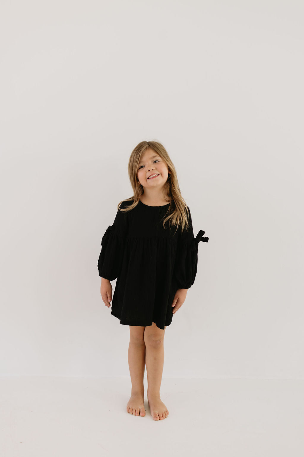 A young girl with long hair stands barefoot against a plain white background, wearing the stylish Aria dress by forever french baby. She smiles directly at the camera, exuding charm as timeless as her outfit.