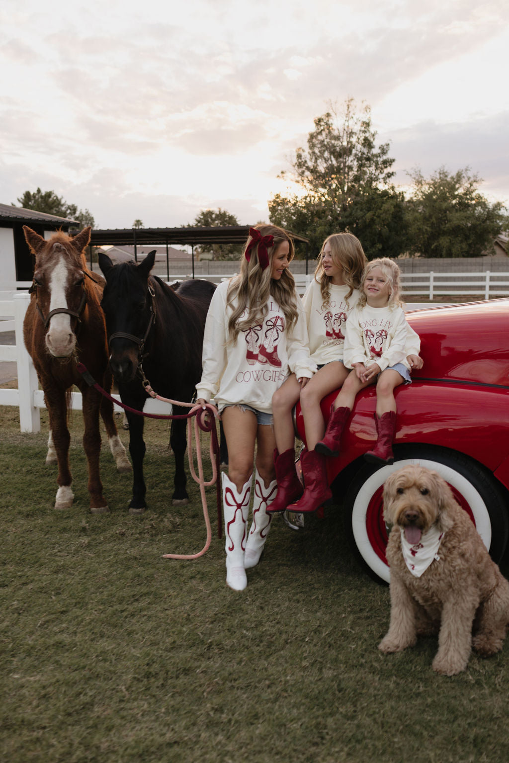 A woman with two children sits on a red car near a brown and a black horse. They wear matching "Cowgirl" outfits made from bamboo viscose, paired with red boots. Nearby, a fluffy brown dog sports one of the latest holiday-themed pet bandanas from lolo webb's Bamboo Bandana collection, specifically the Cowgirl Christmas design. A white fence and trees can be seen in the background.