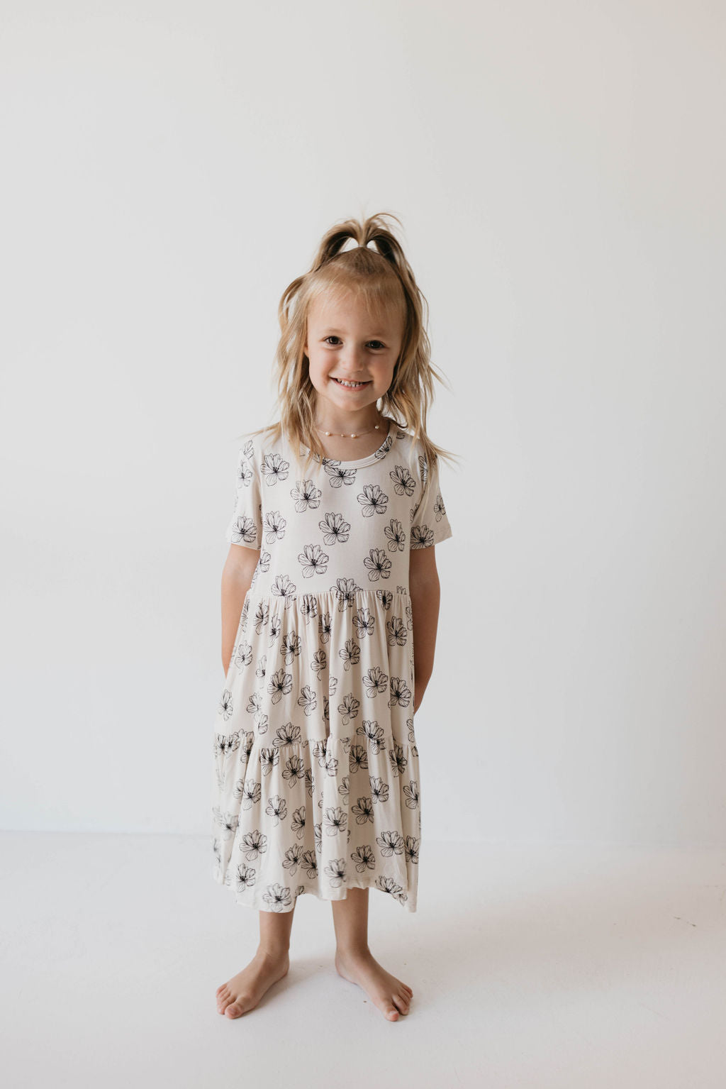 A young girl stands barefoot against a plain white background, smiling at the camera. She is wearing the "Multi Tier Bamboo Dress | Desert Bloom" by forever french baby, a sustainable bamboo fabric dress with short sleeves and black floral designs. Her blonde hair is styled half-up in a ponytail while the rest hangs loose, and her hands are placed behind her back.