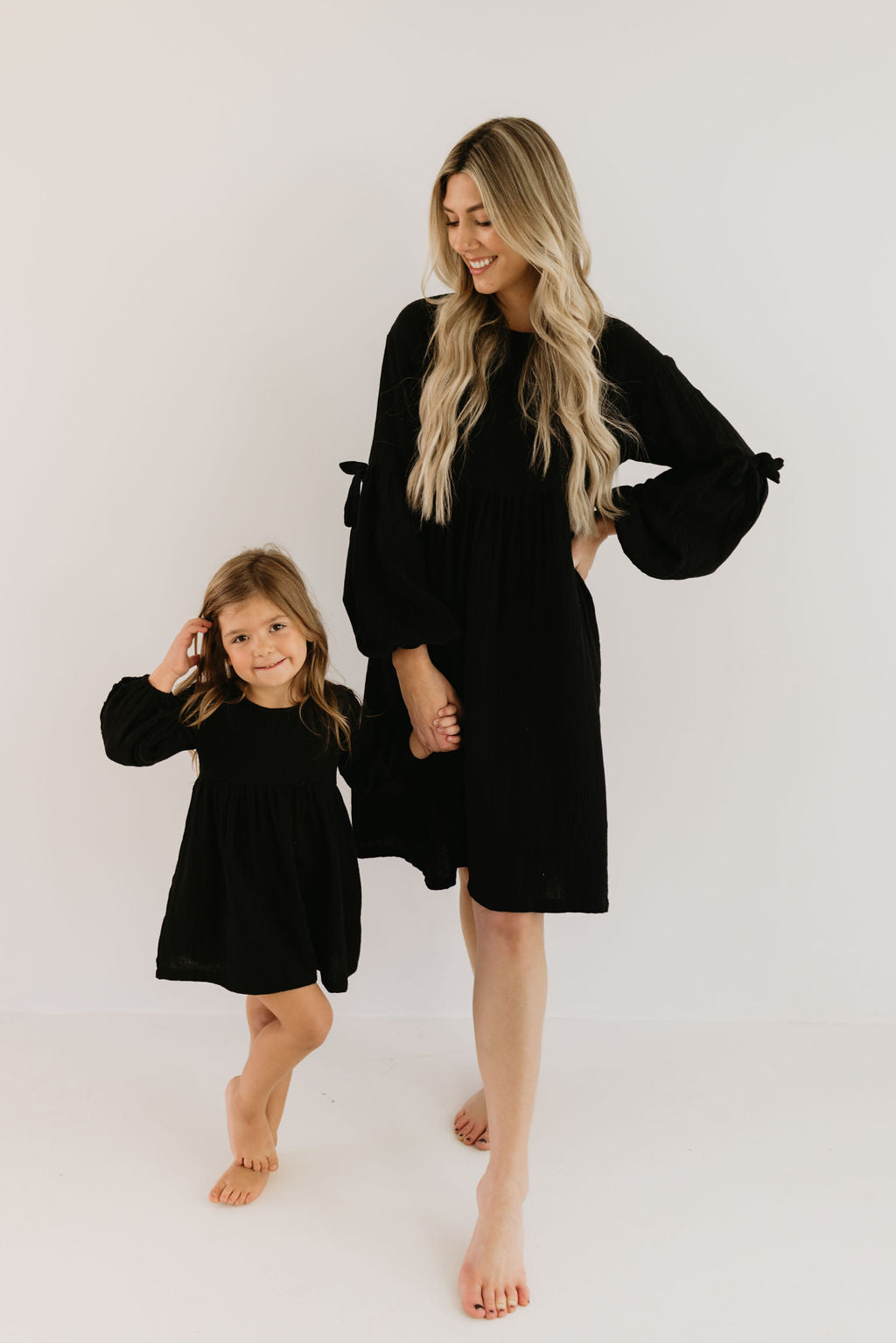 A woman in a forever french baby Aria dress stands barefoot with a young girl, both in matching black dresses, against a neutral backdrop. They hold hands as the girl playfully poses and smiles up at her.