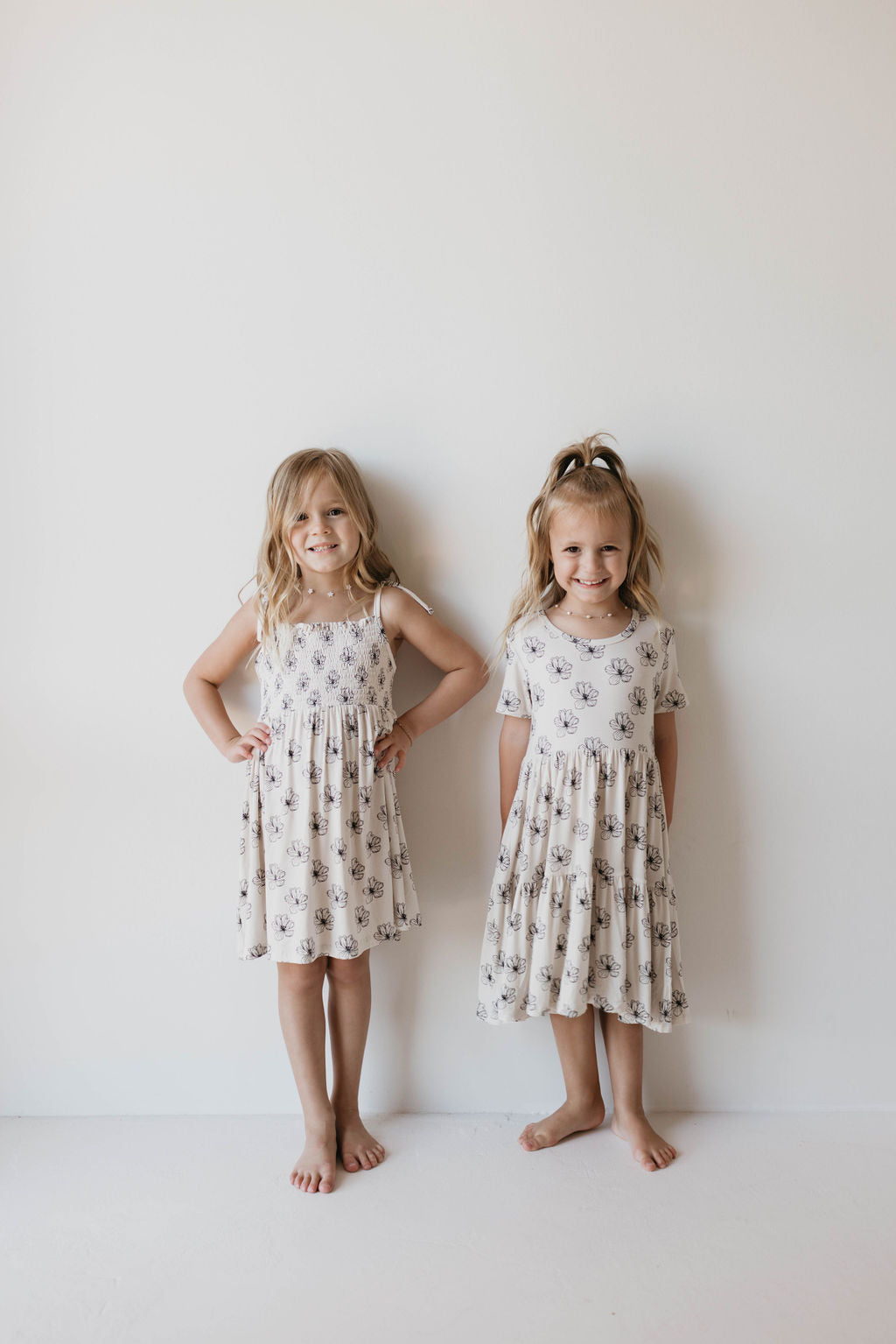 Two young, fashion-forward girls stand side by side against a white wall, both wearing the light-colored and floral-patterned Bamboo Tie Top Dress in Desert Bloom from forever french baby. They are barefoot and smiling, with their arms at their sides. One girl has her hands on her hips, while the other has her hands by her sides.