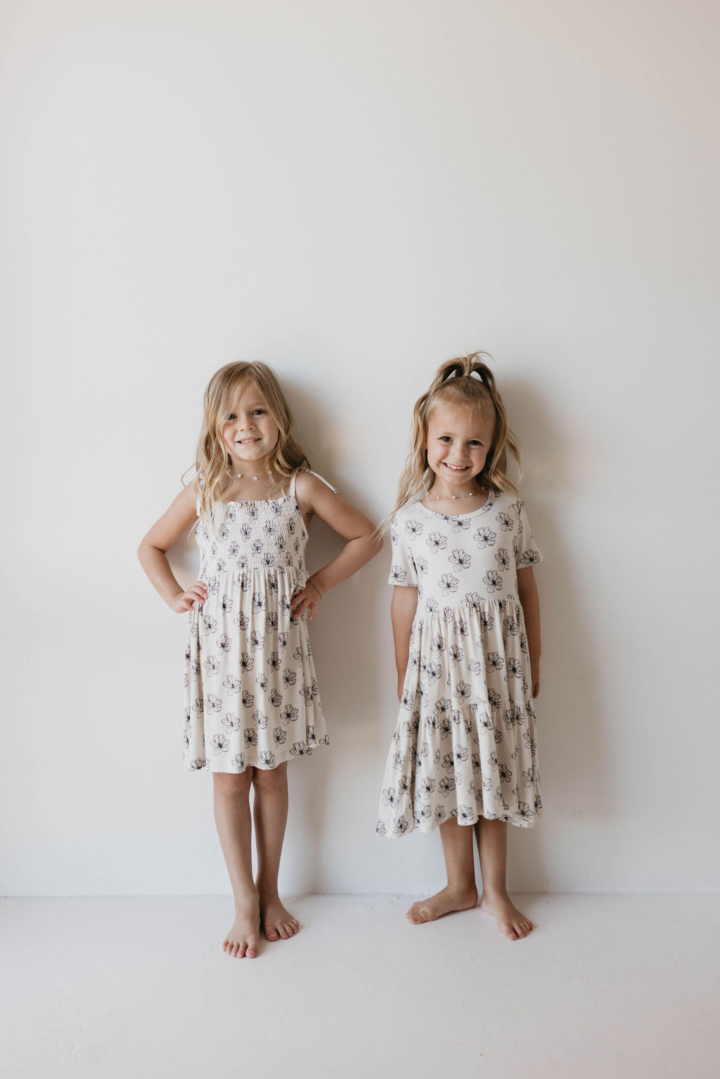 Two young girls stand barefoot against a white wall, each wearing a "Multi Tier Bamboo Dress | Desert Bloom" from forever french baby. The dress features a floral pattern and is made of sustainable bamboo fabric. The girl on the left has her hands on her hips, while the girl on the right has one arm relaxed by her side and a ponytail.