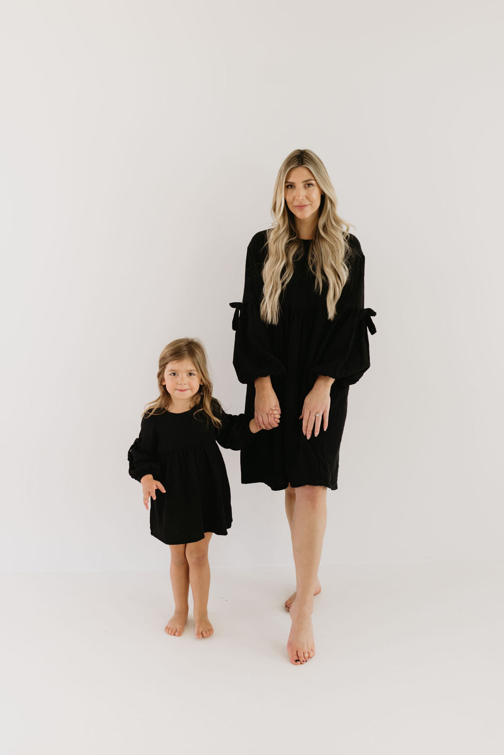 A woman and a young girl, both with long hair, stand barefoot on a white background. They wear matching black "Aria" dresses from forever french baby. The woman holds the girl's hand as they look forward, embodying unity and style.