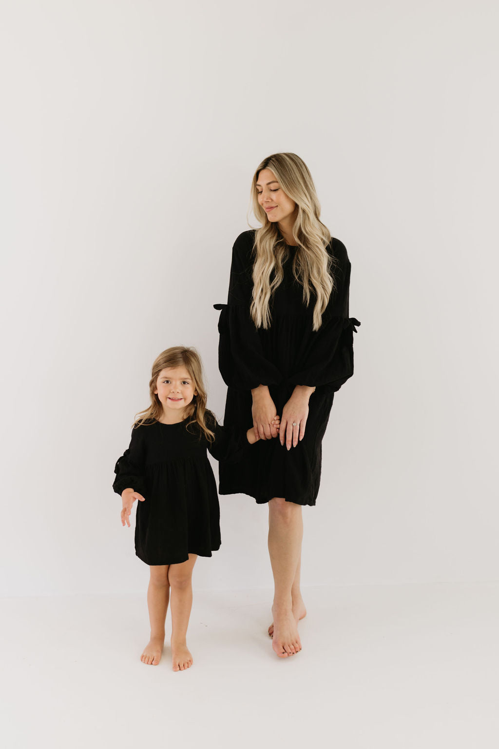 A woman and a young girl stand barefoot against a plain white background, both dressed in stylish black attire. The woman's long blonde hair adds elegance, while the young girl beams at the camera wearing the Aria dress by forever french baby, showcasing their coordinated look.