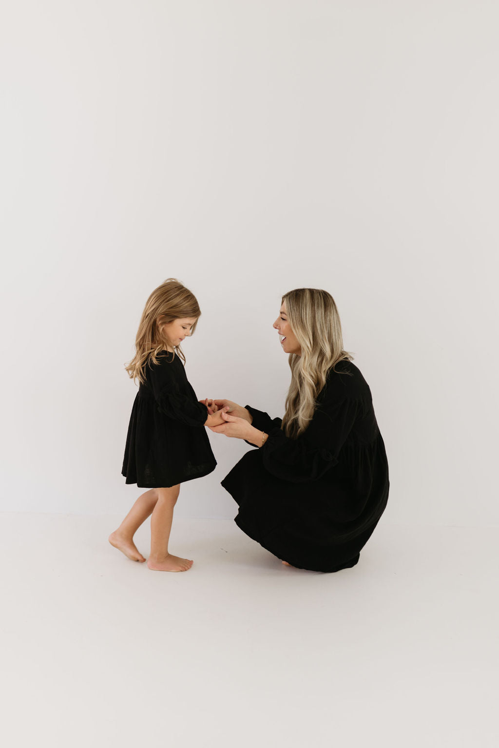 A woman crouches down and smiles at a young girl, both elegantly dressed in matching black Aria dresses from the forever french baby collection, set against a simple, light background. The girl gazes down slightly as the woman affectionately holds her hands.