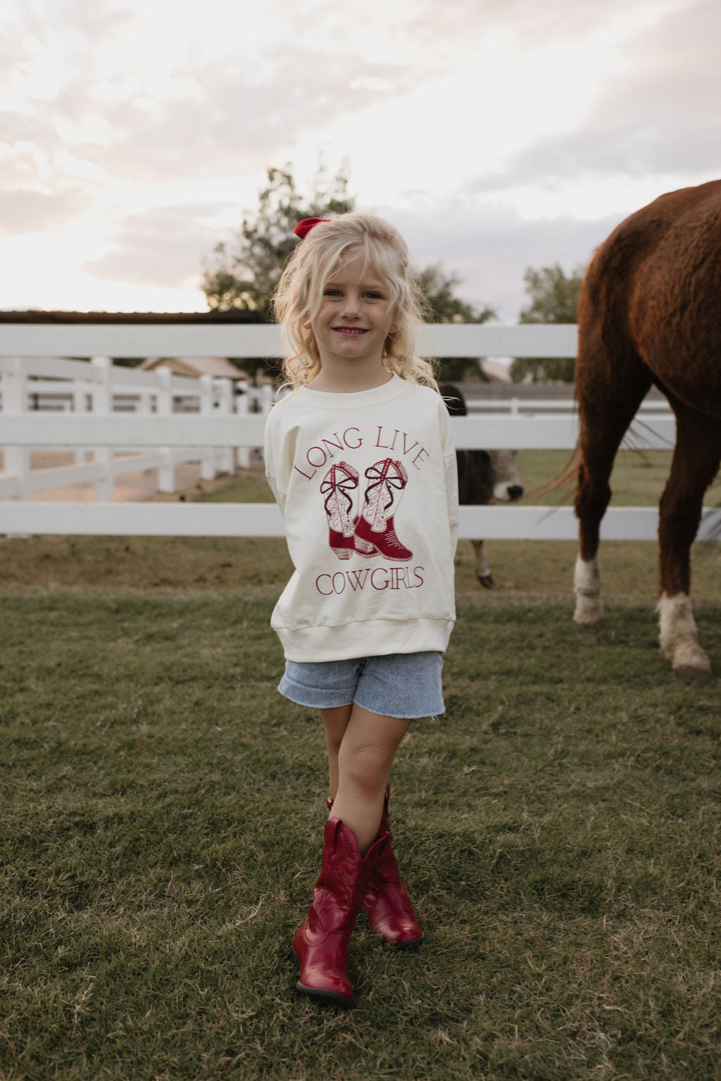 A young girl with blonde hair stands on grass wearing red boots, denim shorts, and a cozy "Long Live Cowgirls" crewneck from lolo webb. A horse grazes behind her next to a white fence under a partly cloudy sky. Check out the matching adult version for fun mother-daughter outings!