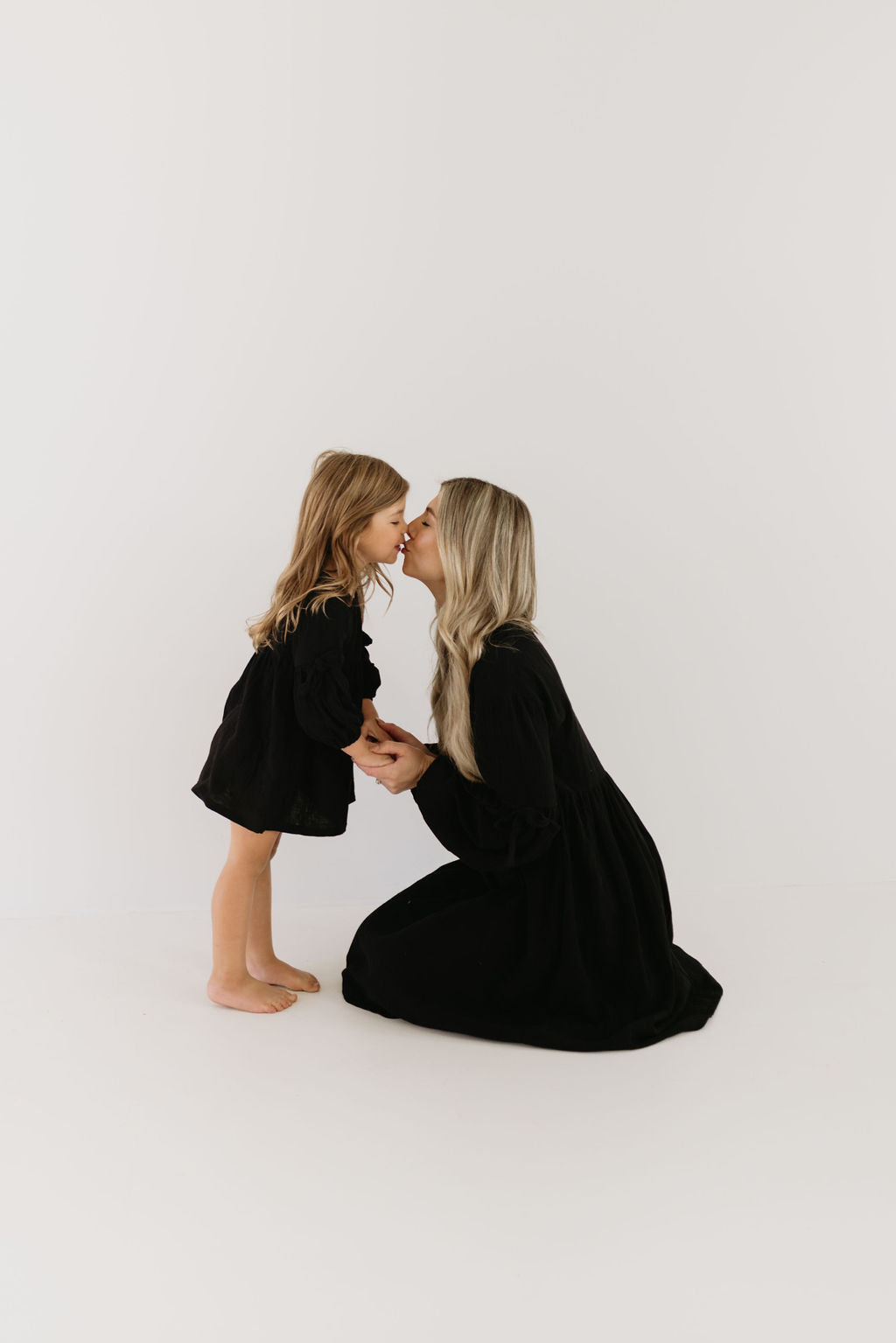 A woman with long blonde hair kneels and kisses a young girl on the forehead. Both wear matching "forever french baby" Child Dress | Aria, showcasing their timeless style against a neutral background.