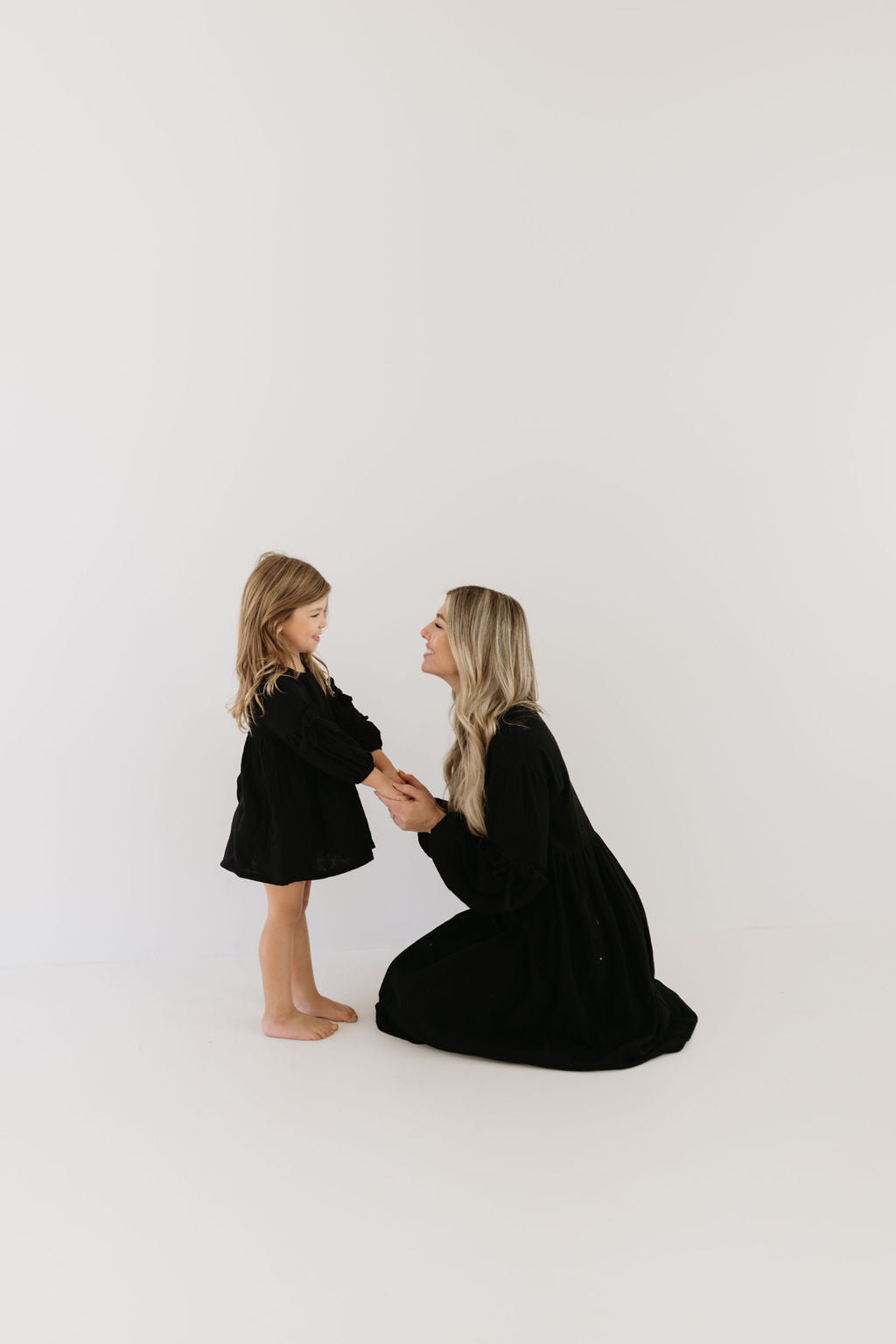 A woman kneels and smiles at a young girl, both wearing matching Aria dresses by forever french baby. The plain white background offers a simple, minimalistic setting, ideal for sharing style care tips.