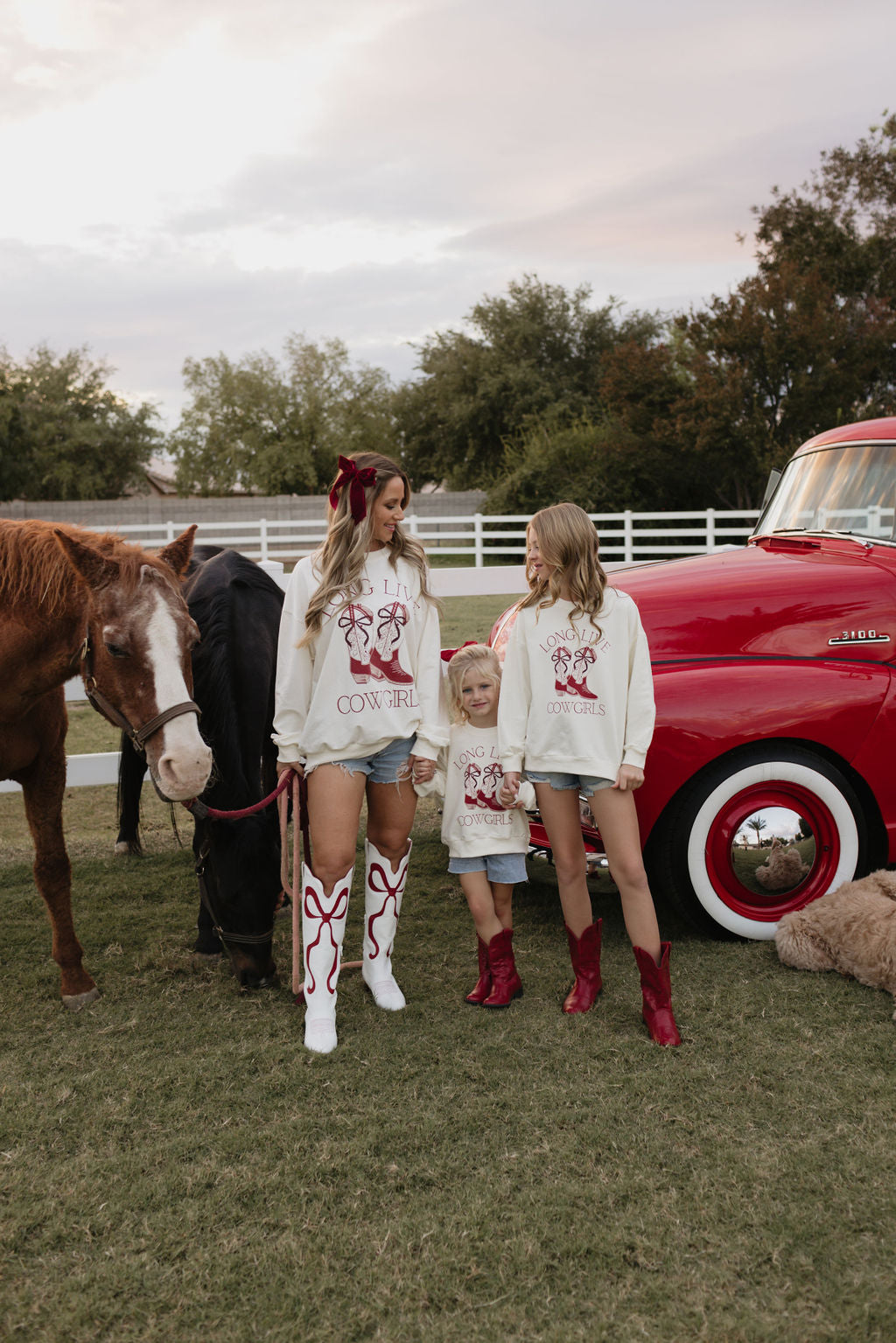 Three people wear lolo webb's "Adult Crewneck | Long Live Cowgirls" sweatshirts and boots, standing on grass with two horses and a vintage red truck. The person on the left holds a horse's reins while the others smile. Trees and a white fence frame the scene, alongside matching children's versions of their outfits.