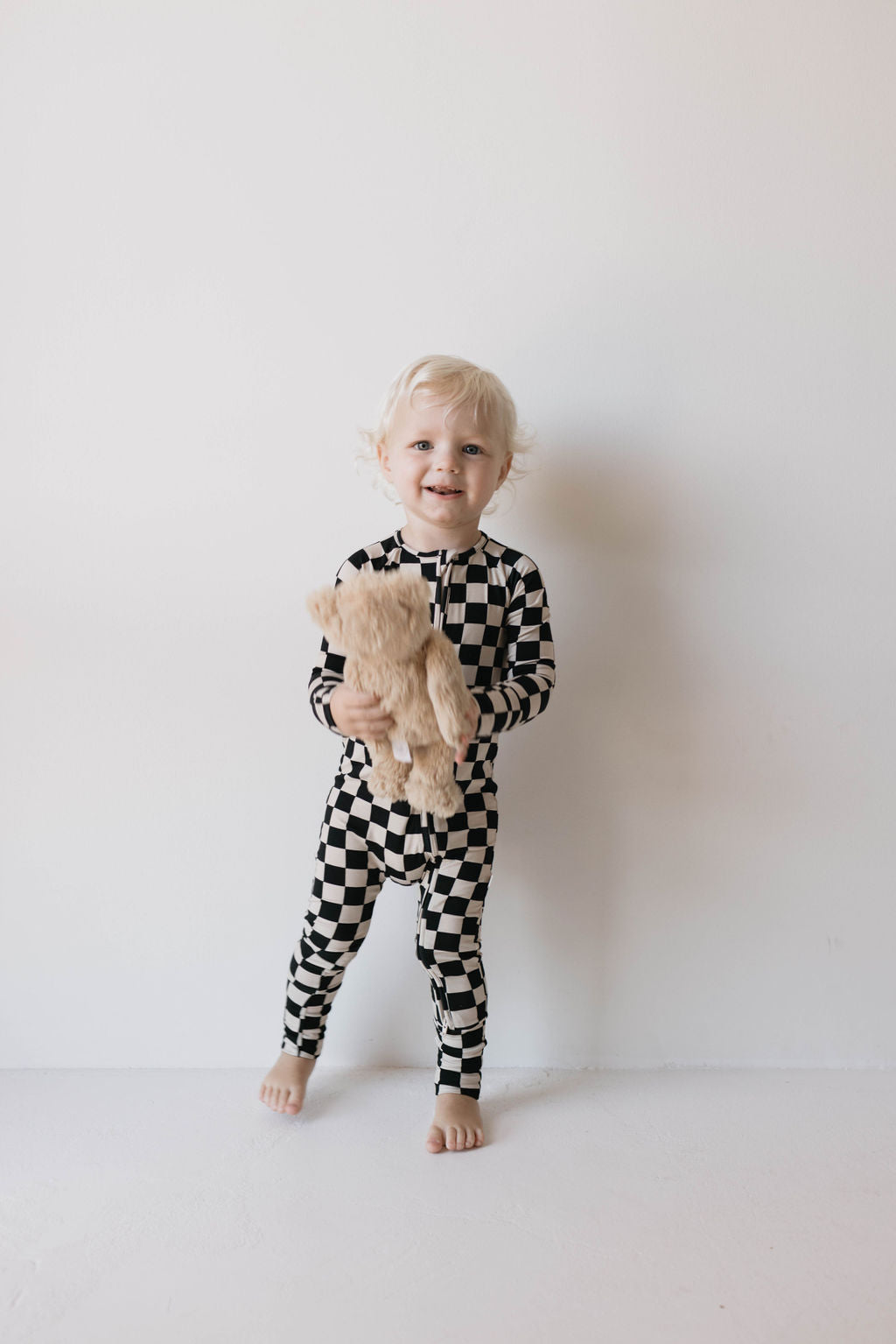 A small child with curly blond hair is standing against a plain white background, wearing forever french baby's Bamboo Zip Pajamas in the Black Checkerboard pattern featuring rollover cuffs. The child is barefoot, holding a light brown teddy bear, and smiling at the camera.
