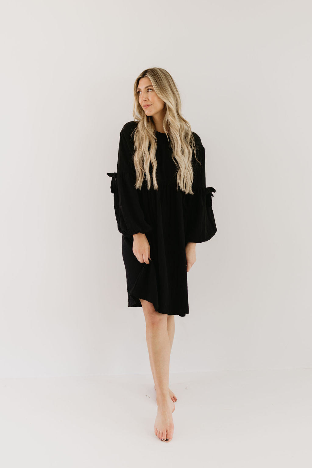 A long-haired blonde woman stands barefoot against a white background, exuding relaxed elegance in a black "Aria" dress by forever french baby. The dress features long, billowy sleeves with tied cuffs. She gazes calmly to the side.