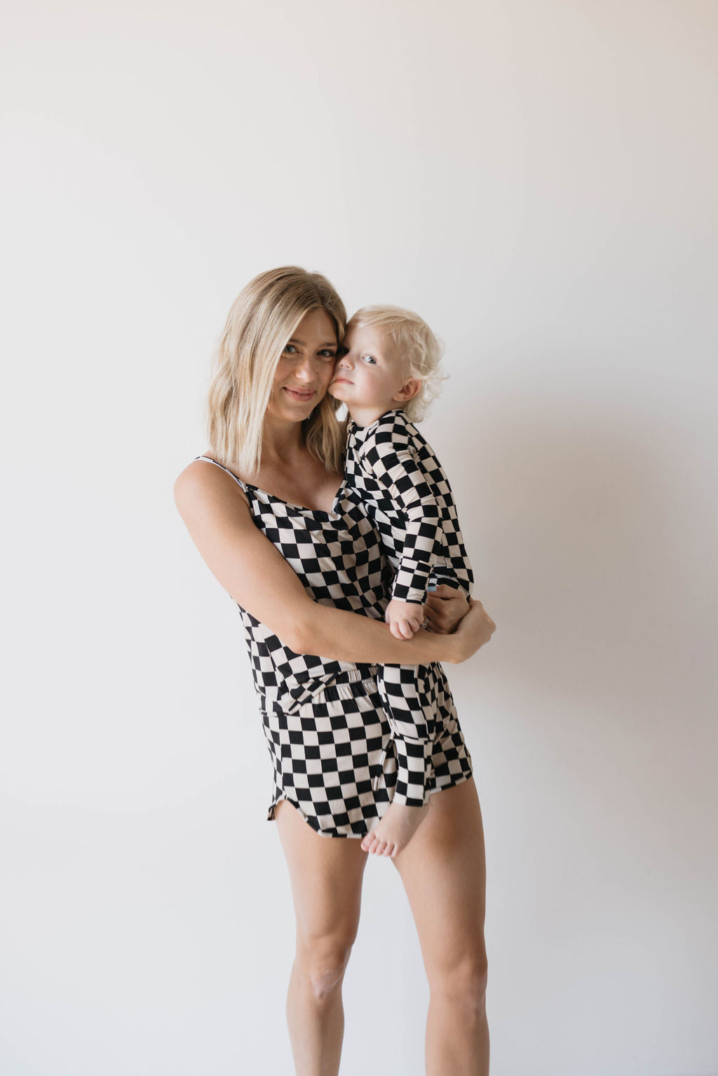 A blonde woman stands smiling and holding a small child. Both are wearing the Forever French Cami Women's Bamboo Set in Black Checkerboard, made from hypo-allergenic bamboo fabric. The child, with light curly hair, is looking off to the side. They are standing against a plain white background.