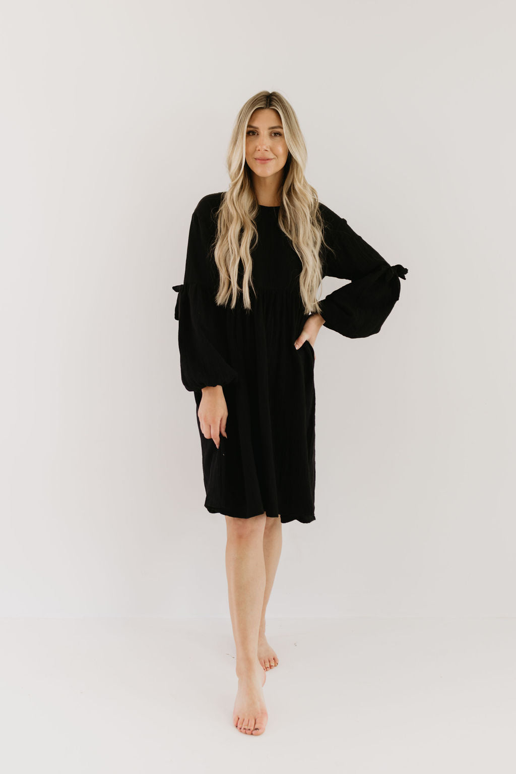 A woman with long blonde hair is elegantly posing barefoot in a black Women's Dress | Aria by forever french baby. Against a plain white backdrop, her attire exudes the effortless style reminiscent of matching rompers, as she smiles gently at the camera.