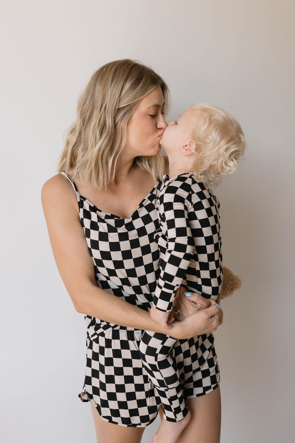 A woman with shoulder-length blonde hair kisses a toddler on the lips. Both are dressed in matching Cami Women's Bamboo Sets from Forever French, featuring a black checkerboard pattern and made from breathable bamboo fabric. The woman cradles the toddler in her arms against a plain background.