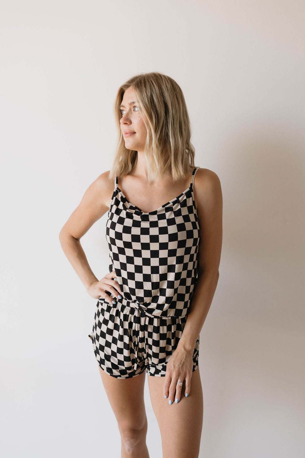 A woman stands against a plain wall wearing the Forever French Cami Women's Bamboo Set in Black Checkerboard. Made from breathable bamboo fabric, the sleeveless romper features a tied waist and complements her shoulder-length blond hair as she gazes to her left with a hand resting on her hip.