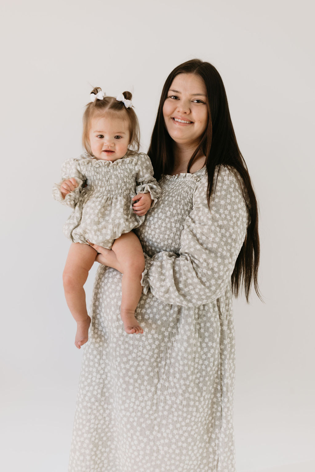 A woman with long, dark hair and a smiling baby both wear forever french baby's Adult Dress in French Gray Floral. The matching outfits highlight their bond, with the baby's small bows adding an adorable touch, as they pose stylishly against a plain white background.