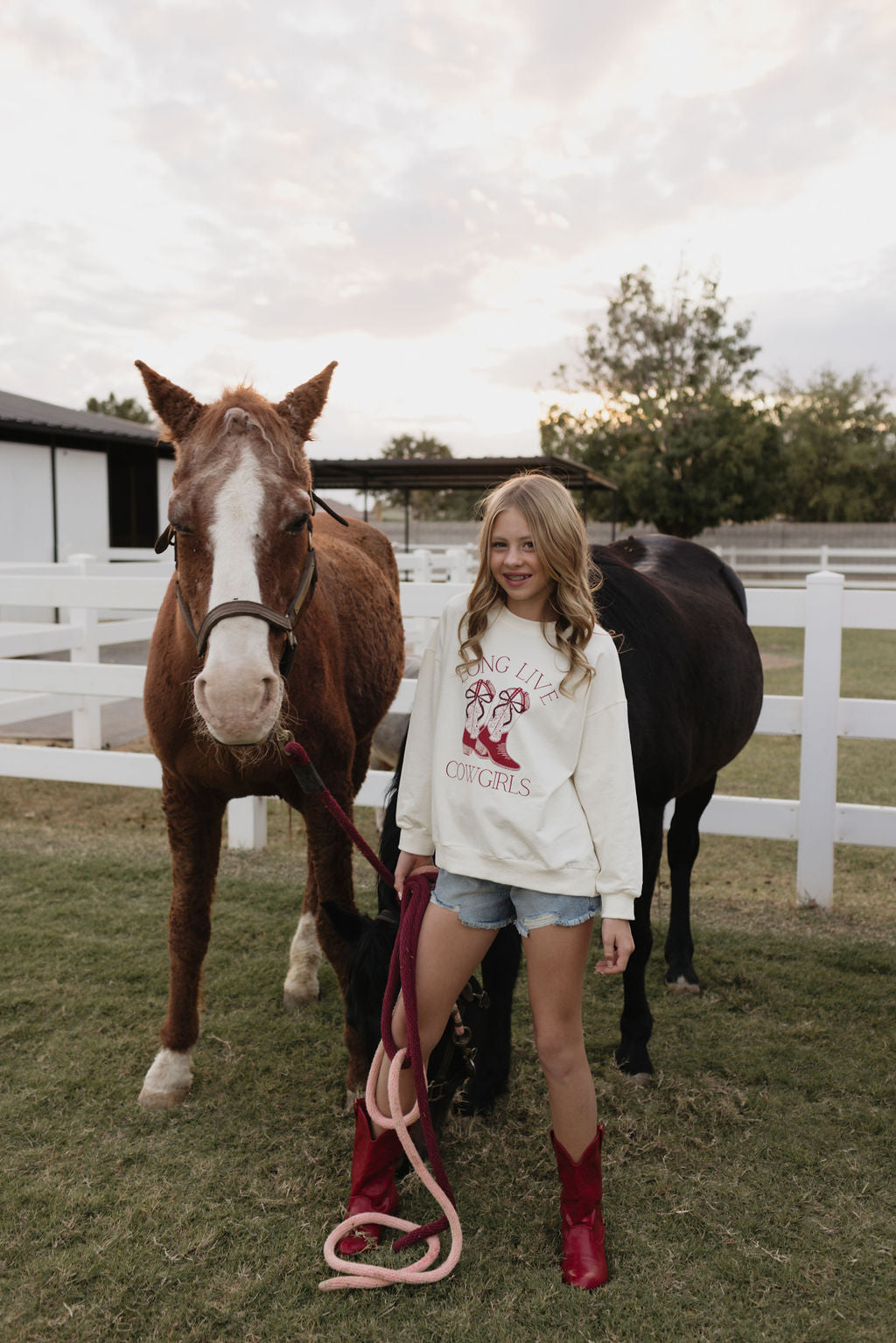 A girl stands in front of two horses in a fenced field, wearing the "Kids Crewneck | Long Live Cowgirls" by lolo webb, paired with denim shorts and red cowboy boots, holding a red lead rope. The partly cloudy sky enhances the relaxed, rural feel.