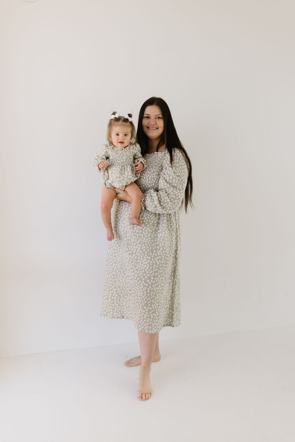 A long-haired woman in an Adult Dress - French Gray Floral by forever french baby holds a baby wearing a matching outfit and small bow against a plain background. The baby, in a smaller size, gazes directly at the camera.