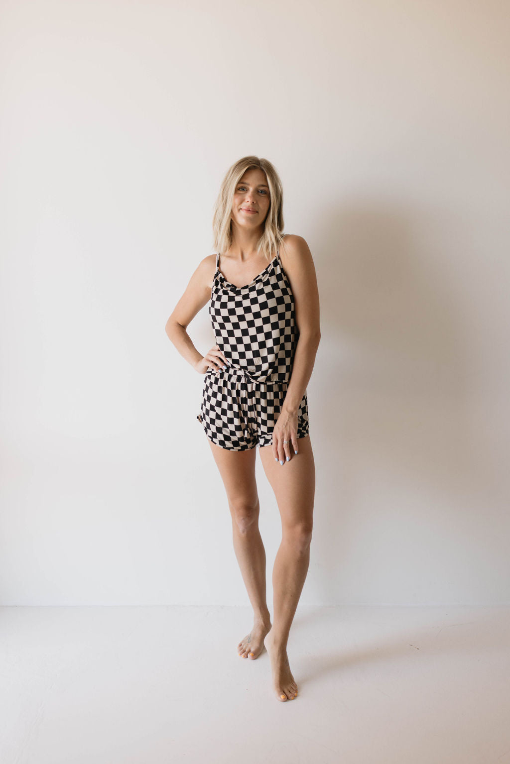 A person with long hair is standing barefoot against a plain background, posing confidently with one hand on their hip. They are wearing the Forever French Cami Women's Bamboo Set in Black Checkerboard, made of breathable bamboo fabric.