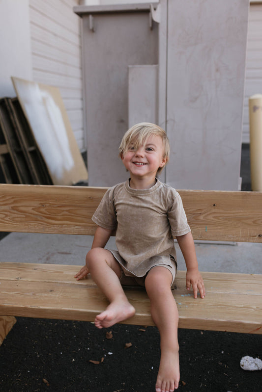 A young child with light hair, dressed in a Vintage Washed Espresso Children's Short Set from forever french baby, sits barefoot on a wooden bench, smiling happily. In the background, there are wooden panels and a building with a gray exterior.