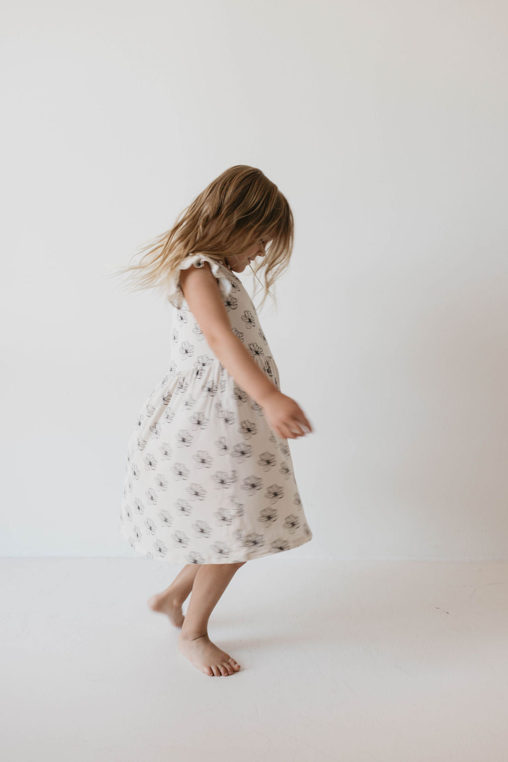 A young girl with long hair wearing a fashion-forward Bamboo Tie Top Dress | Desert Bloom by forever french baby adorned with a floral pattern is spinning around, barefoot, against a plain white background.