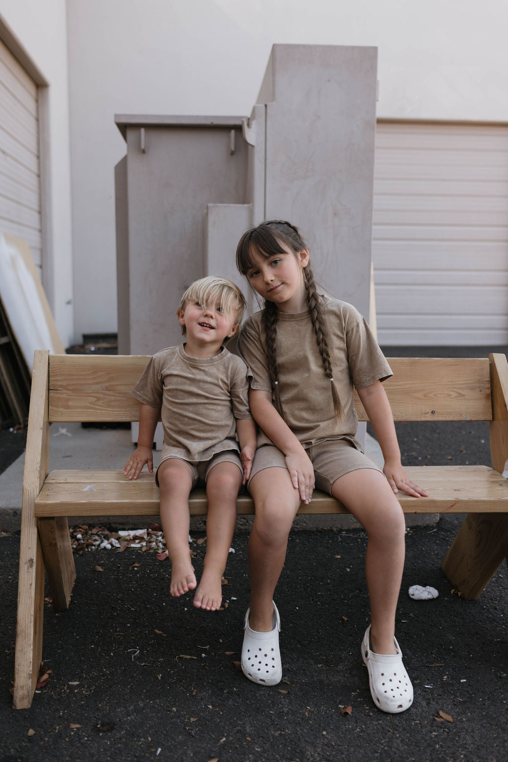 Two children are sitting on a wooden bench. The older child, dressed in a Forever French Baby beige outfit and white Crocs, has long braided hair and smiles slightly at the camera. The younger child, wearing the Children's Short Set in Vintage Washed Espresso by forever french baby, has short blond hair and smiles broadly.