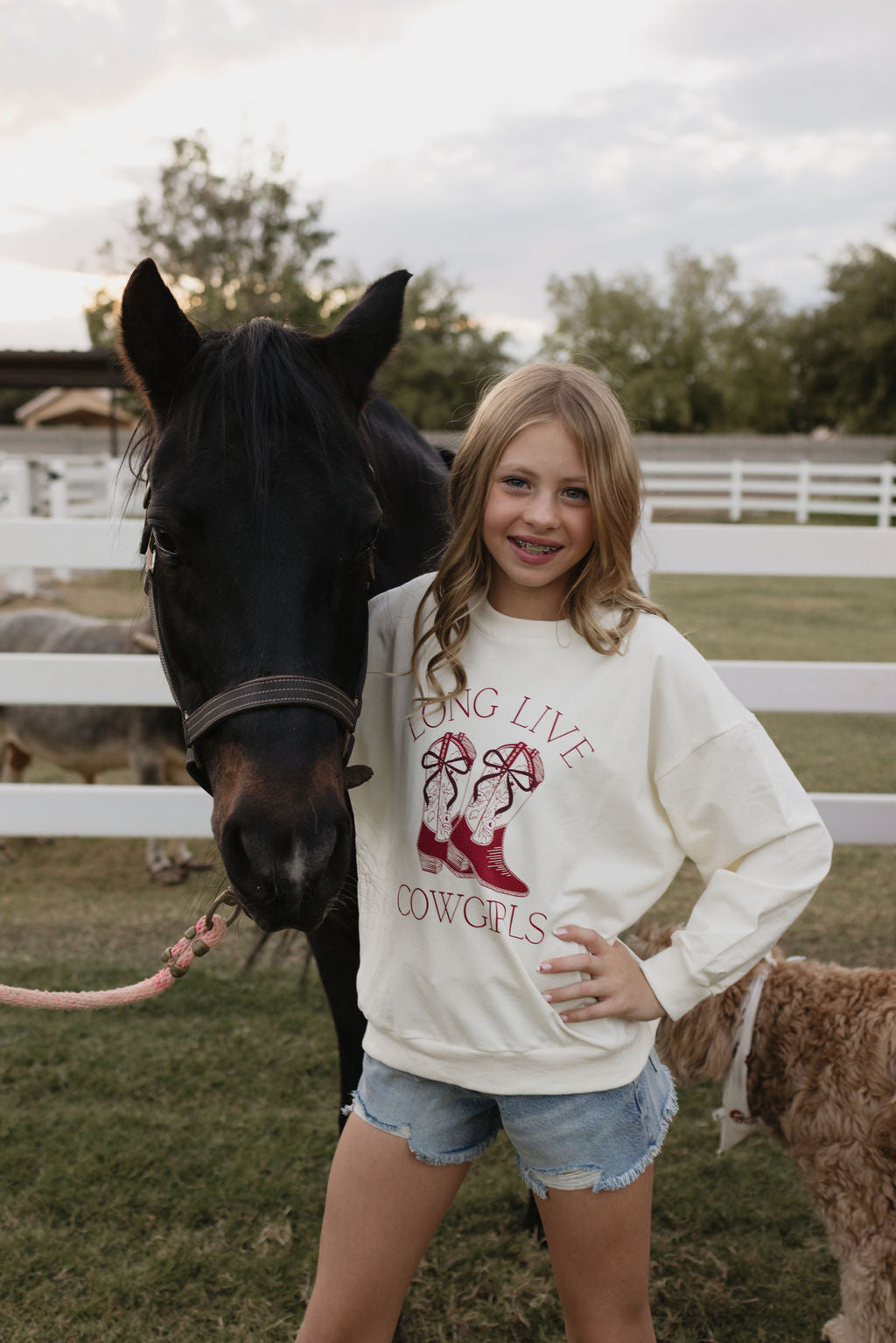 A young girl with long blonde hair beams happily while standing beside a dark brown horse. She is dressed in the lolo webb Kids Crewneck featuring "Long Live Cowgirls" printed on it, complemented by denim shorts. The scene is set outdoors near a white fence, and there's even a matching adult version available in cotton elastane.