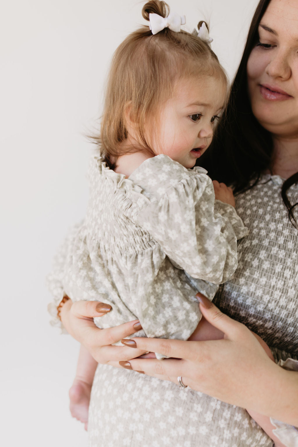 A woman smiles gently against a plain white background, holding a toddler with light brown hair. Both are wearing forever french baby Adult Dress in French Gray Floral, and the toddler is adorned with small bows that complement their matching attire.