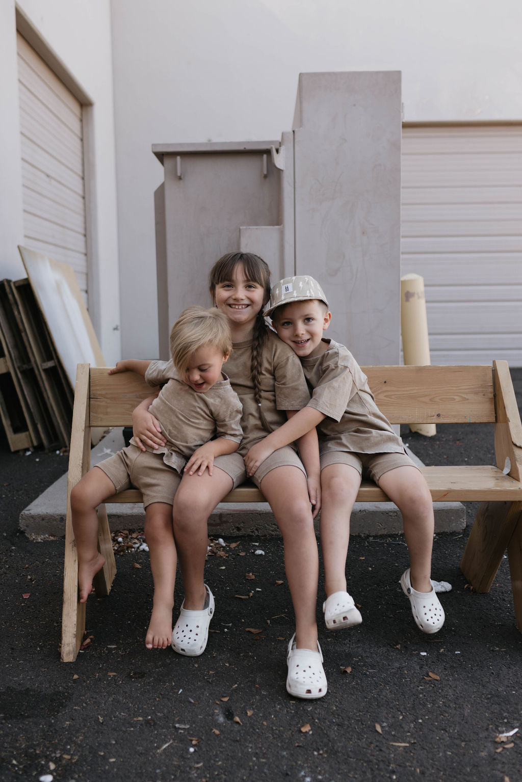 Three children are sitting together on a wooden bench outdoors. The older child in the center is smiling and hugging the younger child on the left, who has blonde hair and is looking down. The child on the right, dressed in a charming Children's Short Set in Vintage Washed Espresso from forever french baby, is smiling at the camera.