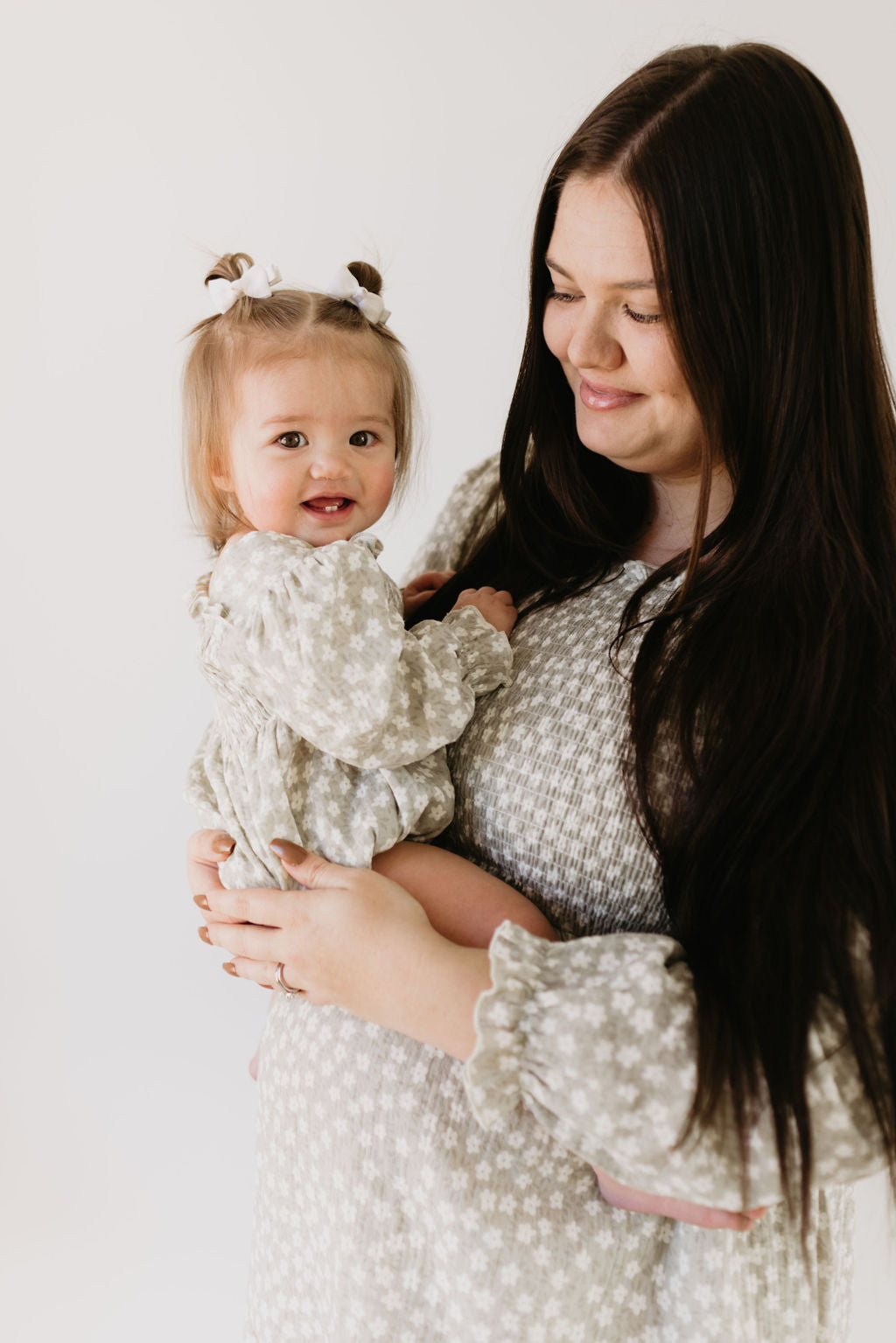 A woman with long dark hair holds a smiling baby, both in matching forever french baby's French Gray Floral outfits. The baby wears a Ruffle Romper accessorized with small bows in her hair, set against an off-white background.