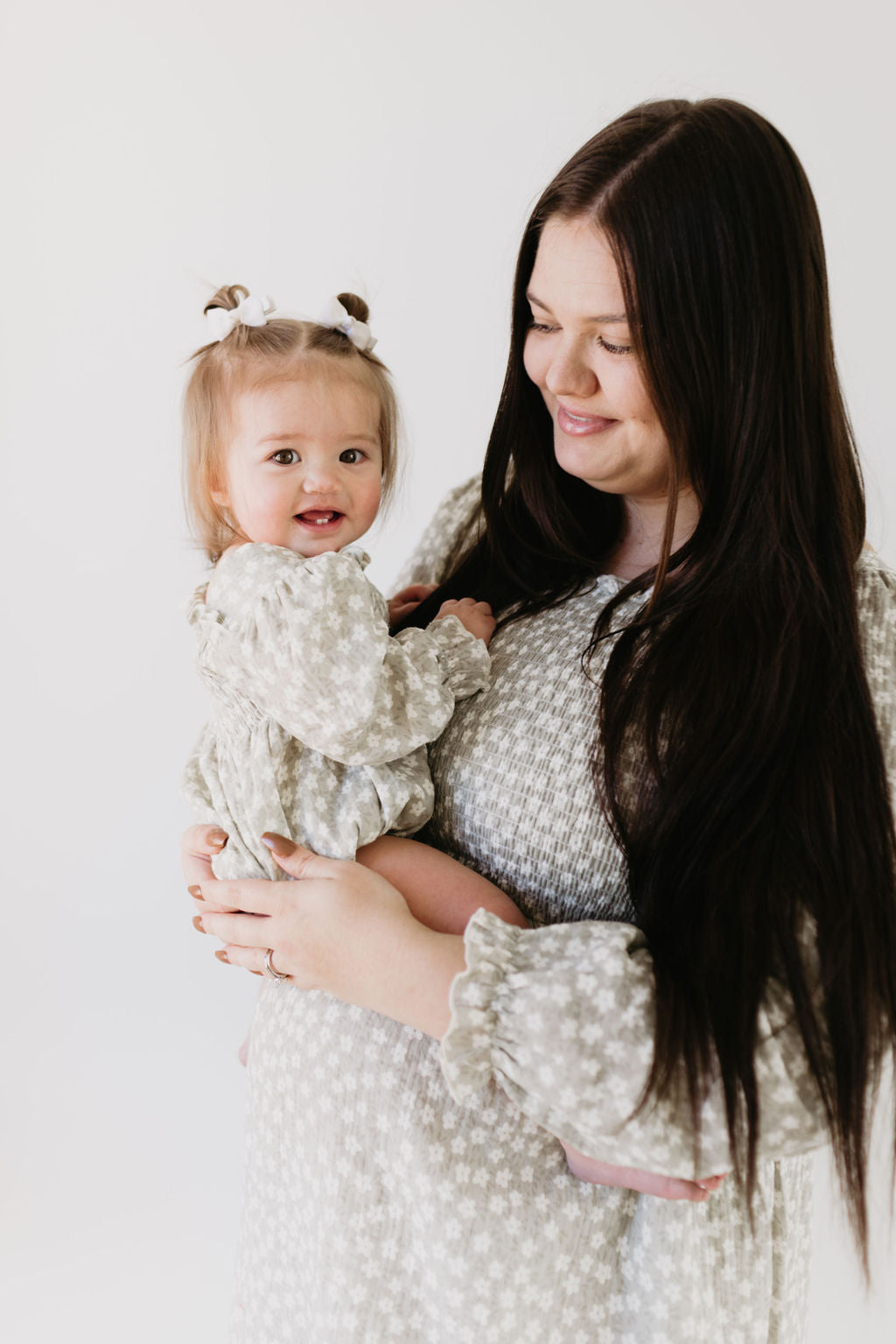 A woman with long dark hair holds a smiling baby girl in a forever french baby's Ruffle Romper in French Gray Floral, with infant-sized pigtails, as they lovingly gaze at each other against a plain background.