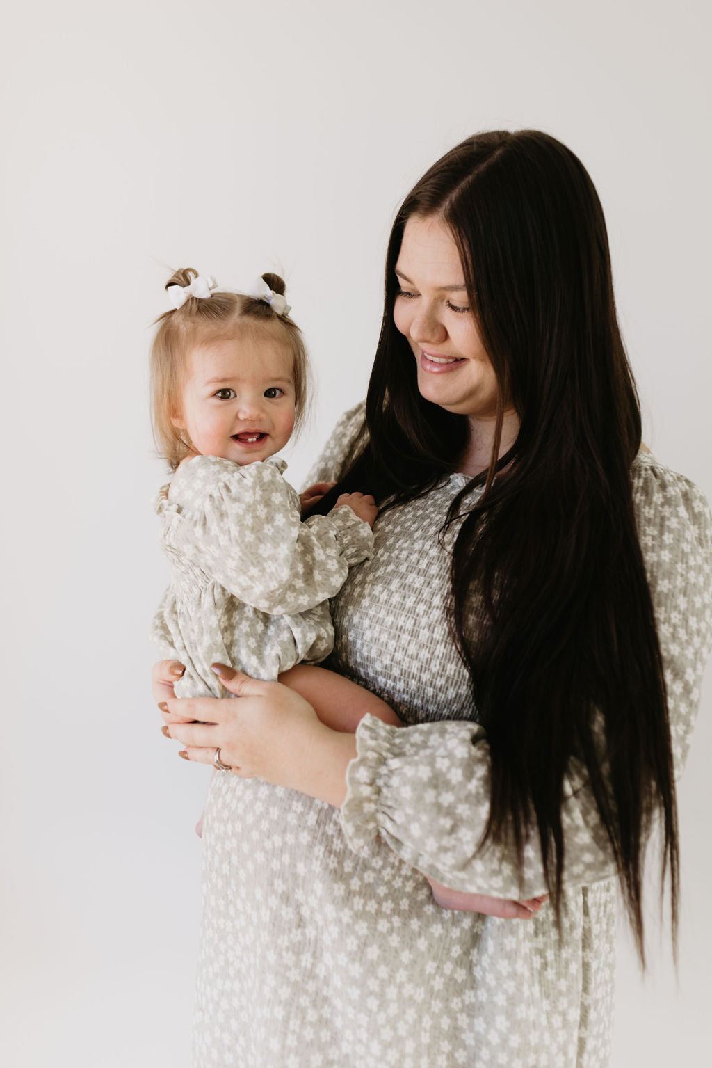 A woman with long dark hair cradles a smiling toddler in pigtails; both wear matching French Gray Floral outfits. The toddler's playful look is enhanced by the forever french baby ruffle romper as the woman gazes at her lovingly against a plain background.