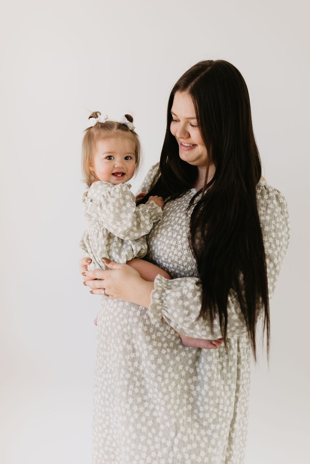 A woman with long dark hair in a French Gray Floral dress holds a smiling toddler. The toddler, wearing a forever french baby Ruffle Romper in French Gray Floral and styled in small pigtails, beams against a plain white background.