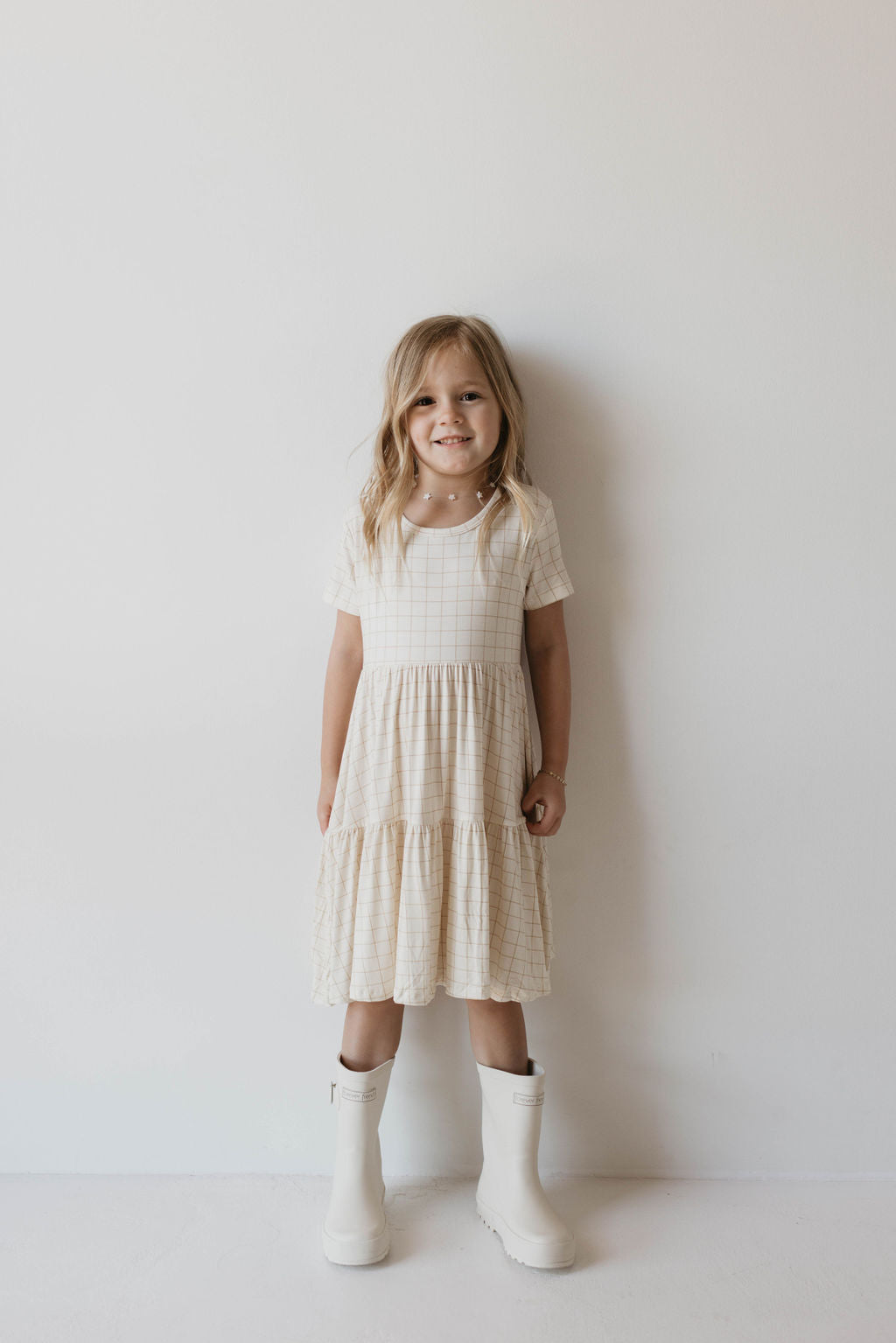 A young girl with long, blonde hair stands smiling against a plain white wall. She is wearing the Multi Tier Bamboo Dress | Golden Grid by forever french baby, featuring a light-colored checkered pattern and tiered skirt, paired with white knee-high boots.