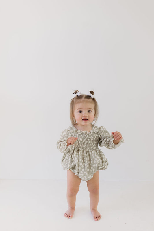 A toddler stands barefoot and curious against a plain white background, dressed in a "Ruffle Romper | French Gray Floral" by forever french baby, with two small ponytails adorned with bows.