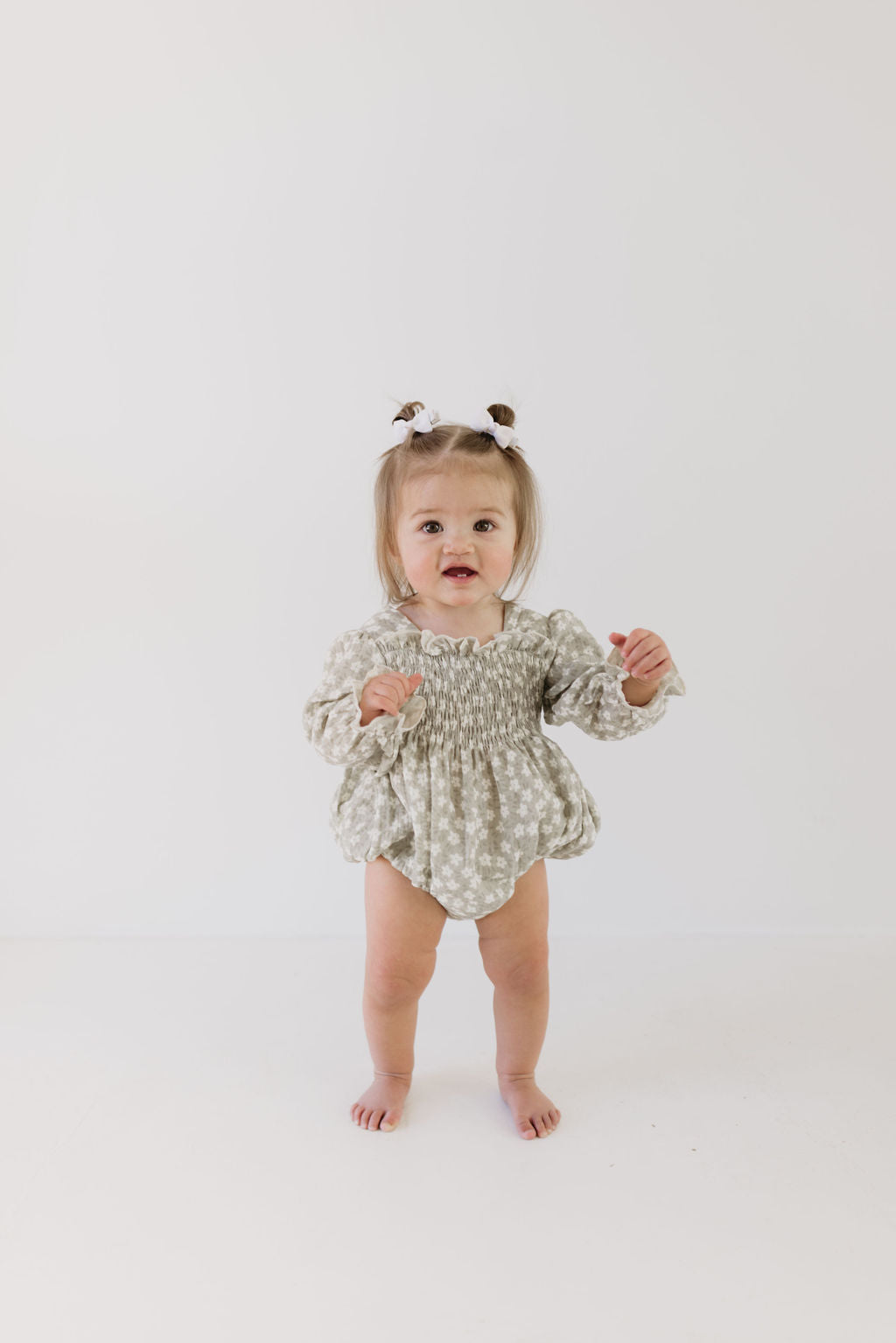 A toddler stands against a plain white background, wearing the Ruffle Romper in French Gray Floral by forever french baby. Her hair is styled with small bows, and she looks forward with a curious expression. Available in infant sizes, it's ideal for adding charm to any little one's wardrobe.