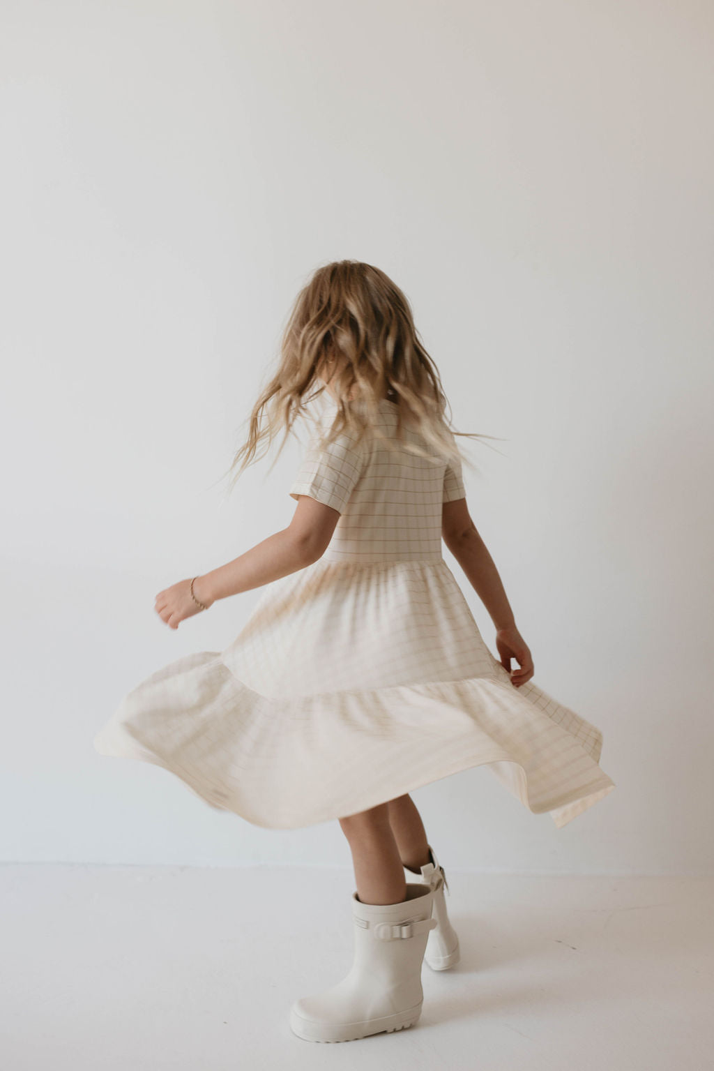 A young girl with wavy light brown hair wears the "Multi Tier Bamboo Dress | Golden Grid" from forever french baby, a cream-colored, short-sleeved dress made from sustainable bamboo fabric featuring a subtle golden grid pattern. She spins around, making her bamboo dress flare out. She pairs the dress with white rubber boots against a plain and light-colored background.